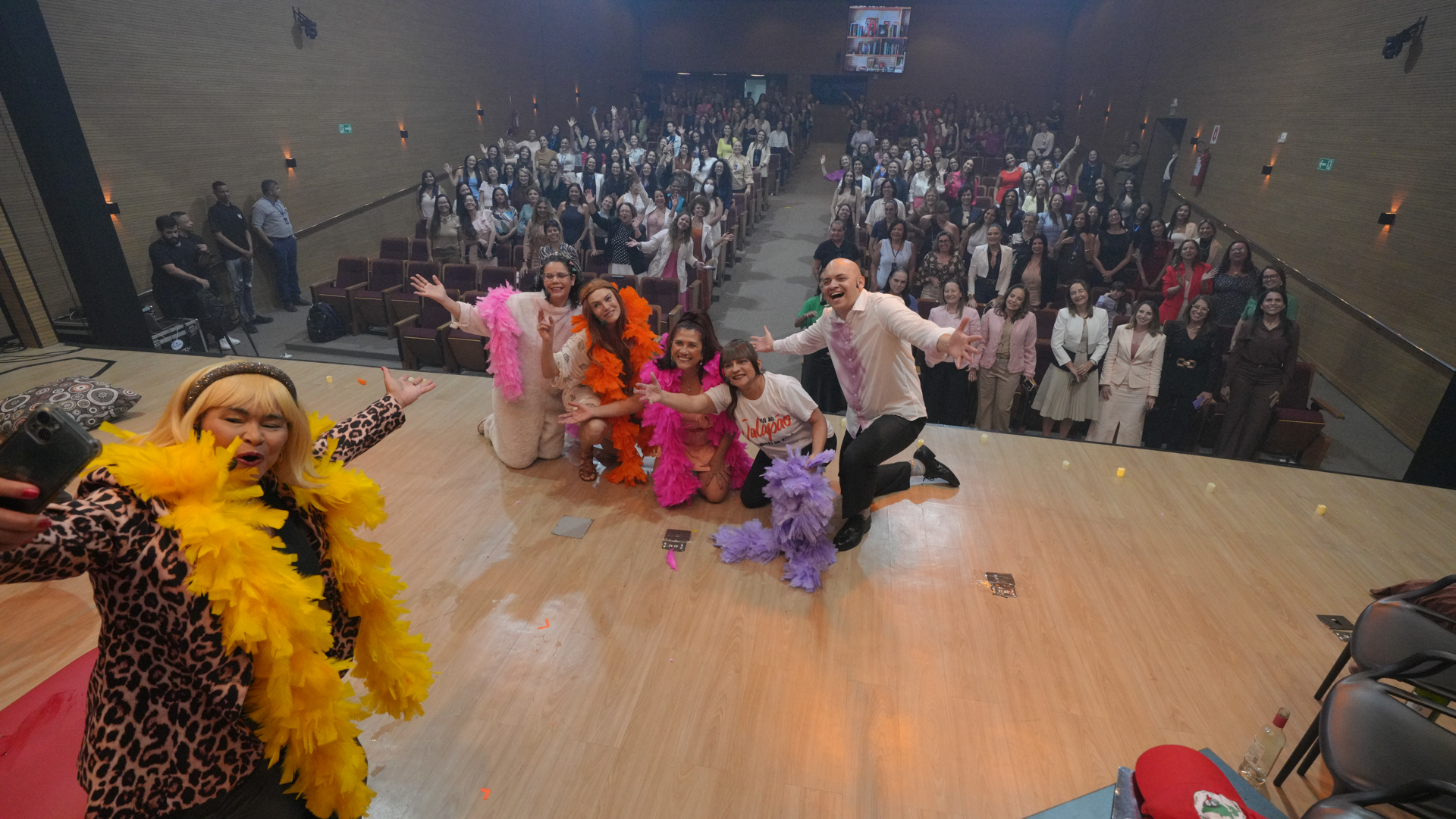 Foto de atores no palco tirando selfie. Ao fundo, plateia em pé, posando para foto