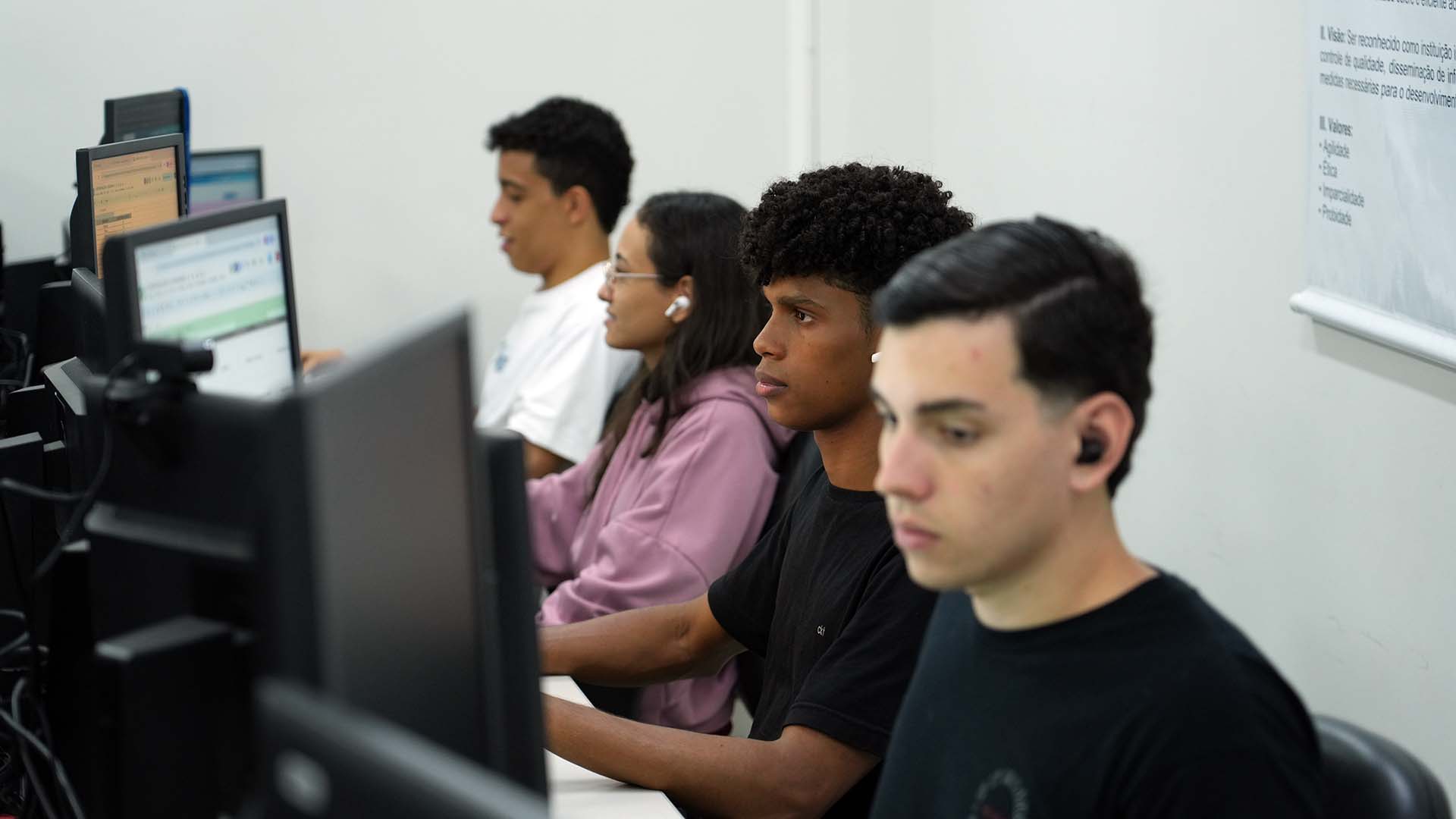 Ambiente de trabalho institucional com servidores do Nacom atuando em computadores de mesa. Em primeiro plano, um servidor, visto de costas, utiliza dois monitores enquanto analisa informações no sistema. Ao lado, uma servidora também trabalha diante do computador. Ao fundo, outros profissionais aparecem em suas estações. Na mesa, há objetos de uso cotidiano, como teclado, mouse, copo plástico e um calendário de mesa.