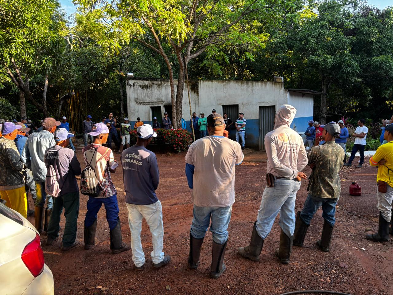 Grupo de trabalhadores participa de uma reunião ao ar livre no Projeto Amigos do Meio Ambiente (AMA), em Porto Nacional. Os participantes formam um círculo em área aberta, cercada por árvores e vegetação, enquanto acompanham orientações durante a atividade. A maioria veste roupas de trabalho, botas e bonés, característicos de atividades operacionais. Ao fundo, uma pequena construção e canteiros com plantas compõem o ambiente.