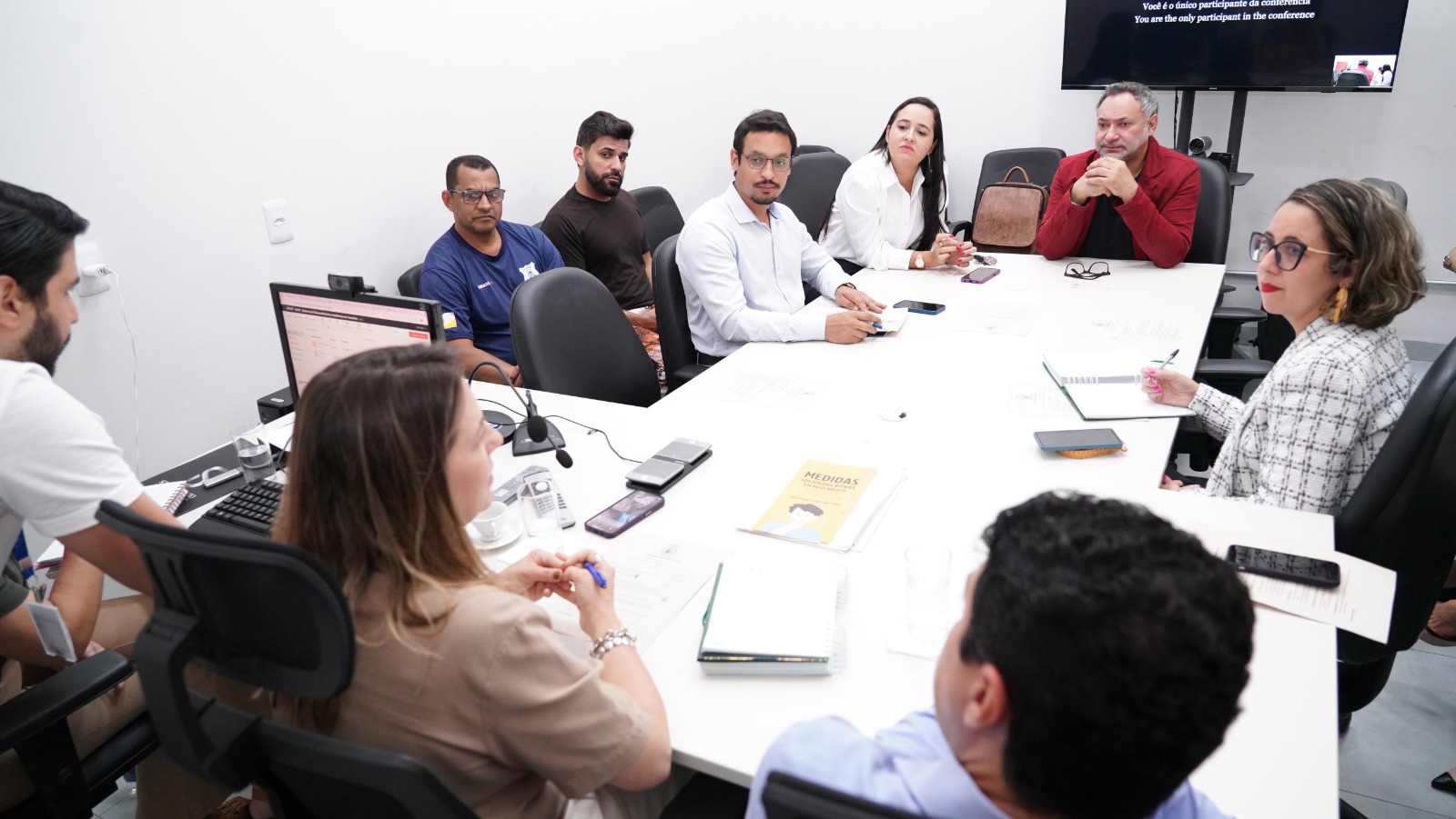 Participantes de reunião institucional sentados ao redor de uma mesa, com documentos, celulares e notebook, debatendo pautas em sala de reuniões