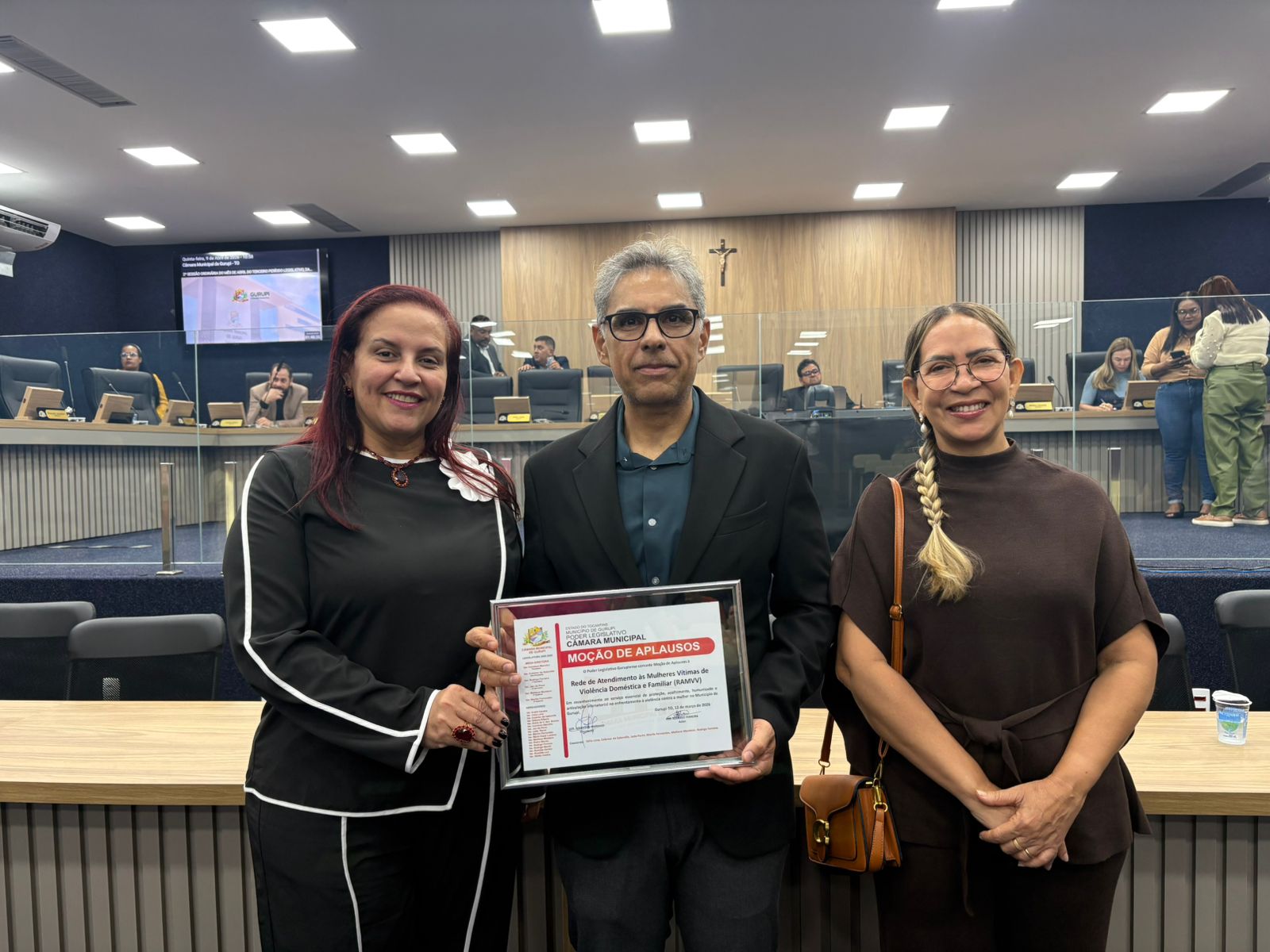 imagem mostra o juiz Jossaner Luna no plenario da Câmara de Gurupi segurando uma placa de homenagem ao lao da secretária Municipal da Mulher e Cidadania, Cristina Donato, Lucinei da Silva Nery Nogueira Luna, analista da defensoria pública