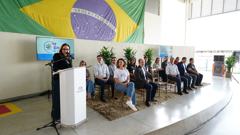 Imagem mostra autoridades no palco, sentadas em cadeiras, e ao fundo a bandeira do brasil aberta na parede