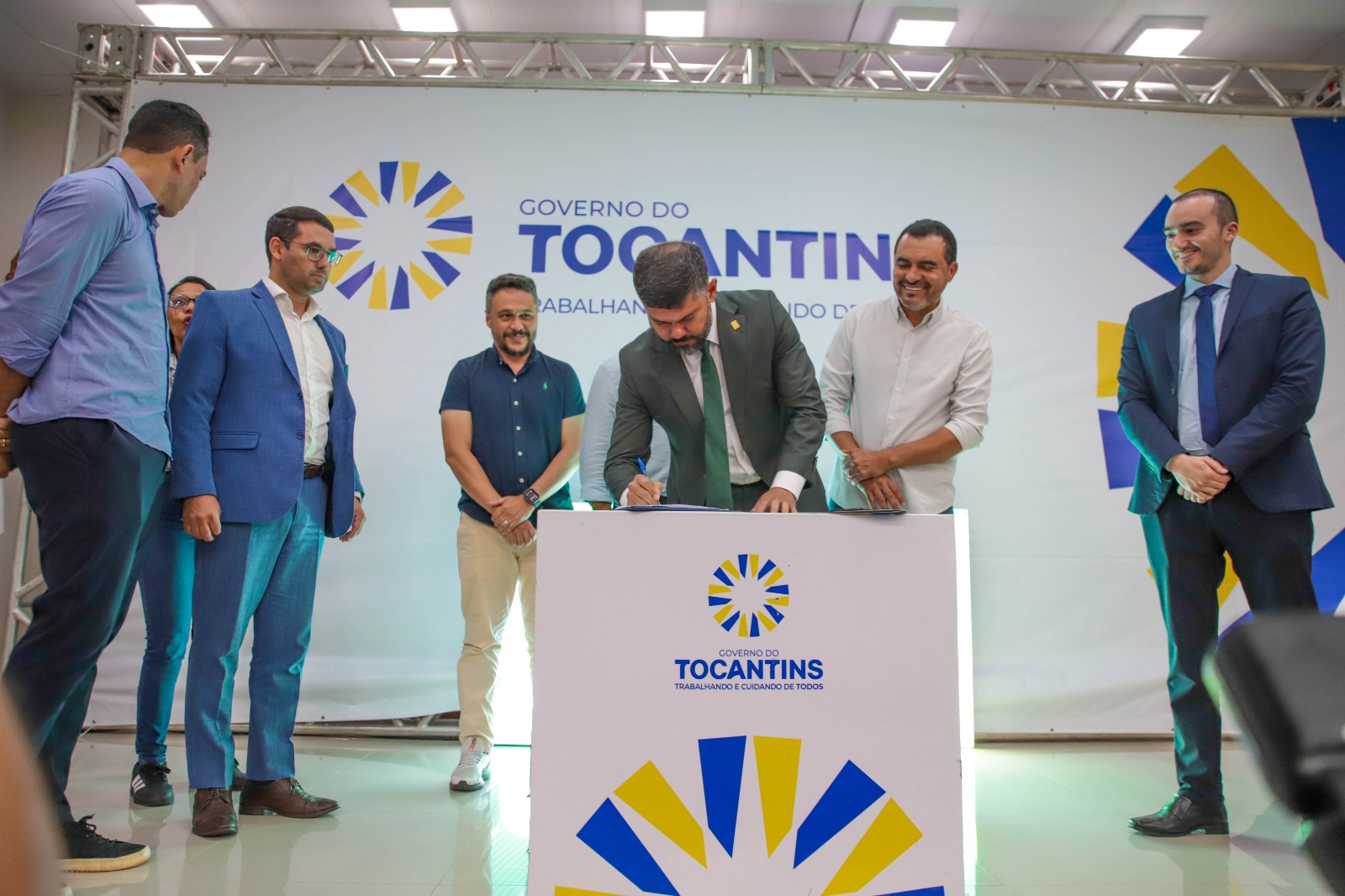 Imagem mostra o juiz Wellington Magalhães assinando convêncio em um púlpito observado por autoridades