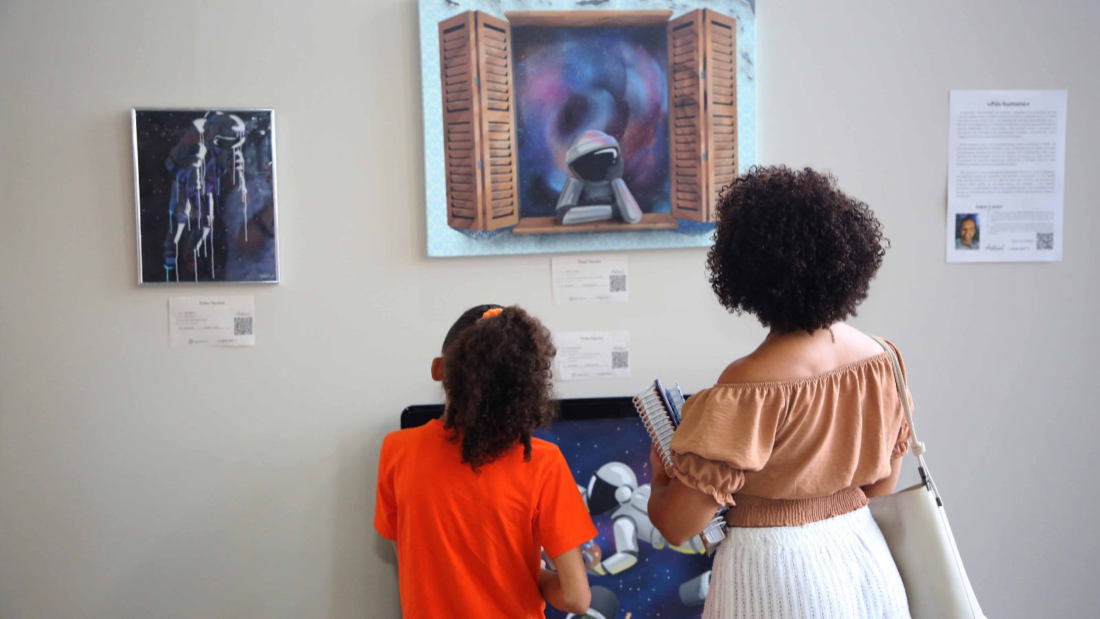 Mulher de cabelo curto, preto e crespo usando saia branca e blusa bege aprecia obras de artes exposta em um mural de madeira ao lado de uma menina de cabelo preto e cacheado com blusa laranja e calça xadrez vermelha