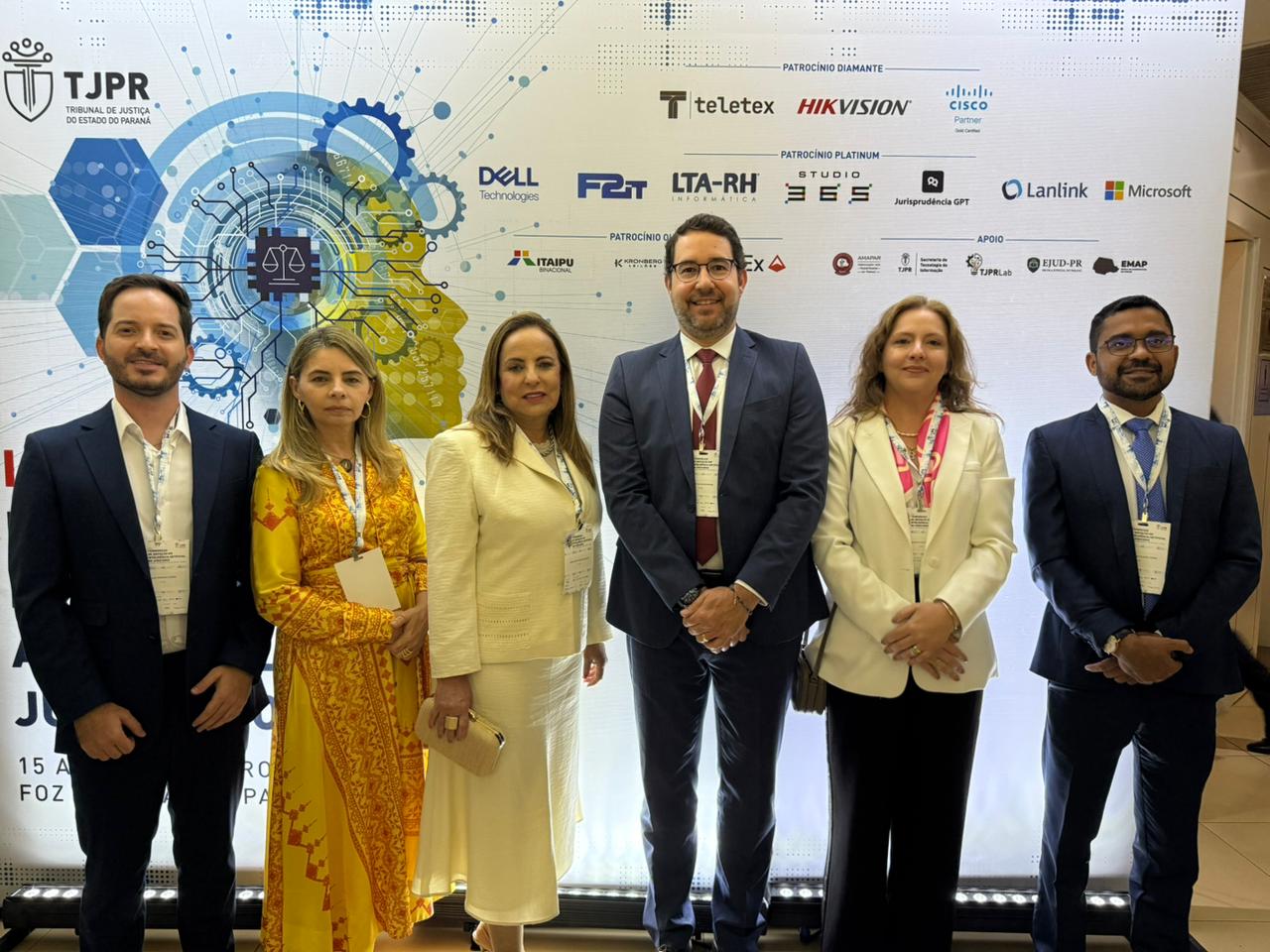 Seis pessoas vestidas formalmente em pé, posando para foto em frente ao painel do evento.