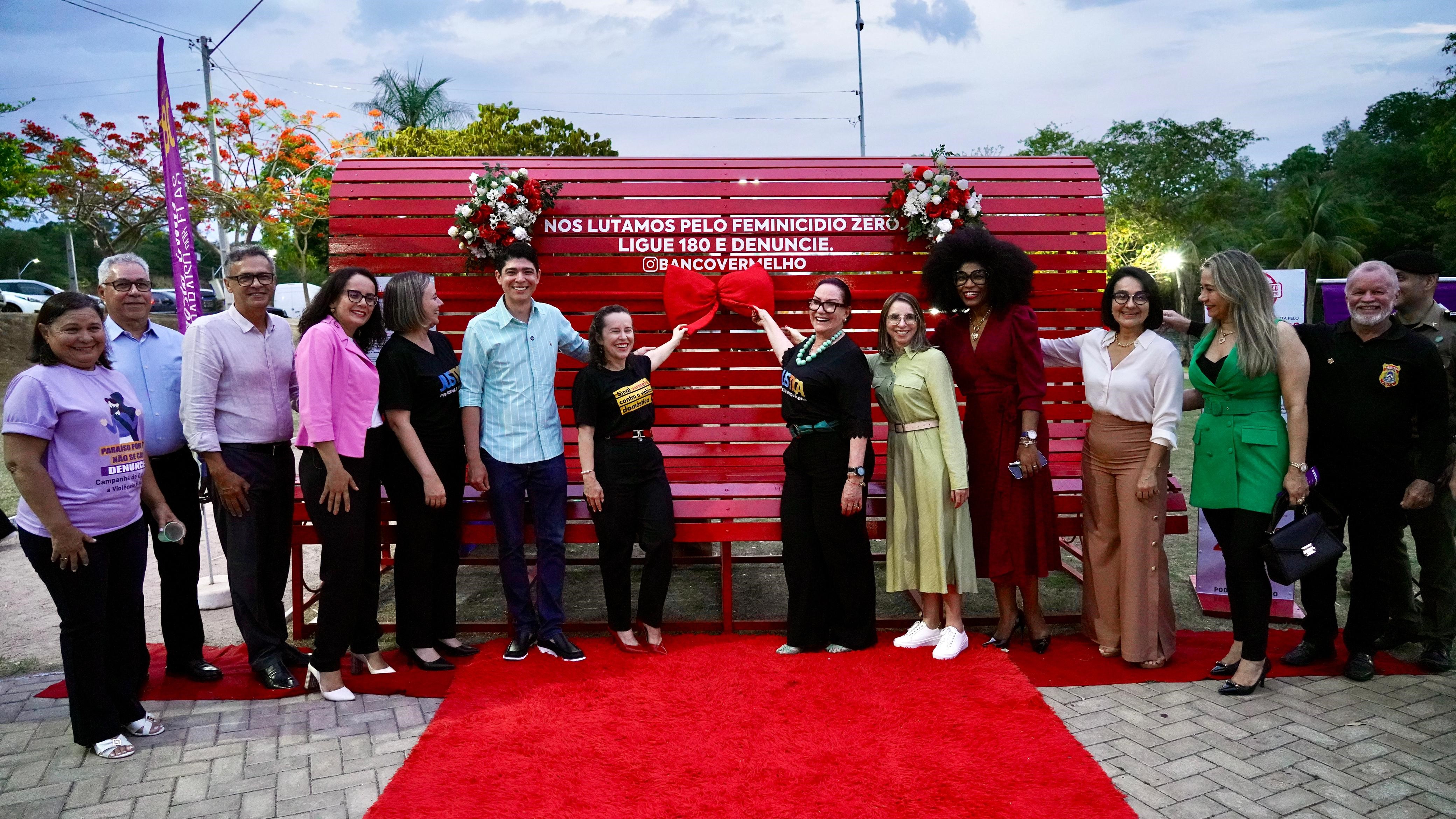 Grupo de pessoas posando em frente a um banco de madeira vermelho com flores nos cantos superiores e com os dizeres: "Nós lutamos pelo feminicídio zero. Ligue 180 e denuncie." Os participantes estão vestidos em trajes formais, ao ar livre e céu levemente nublado.