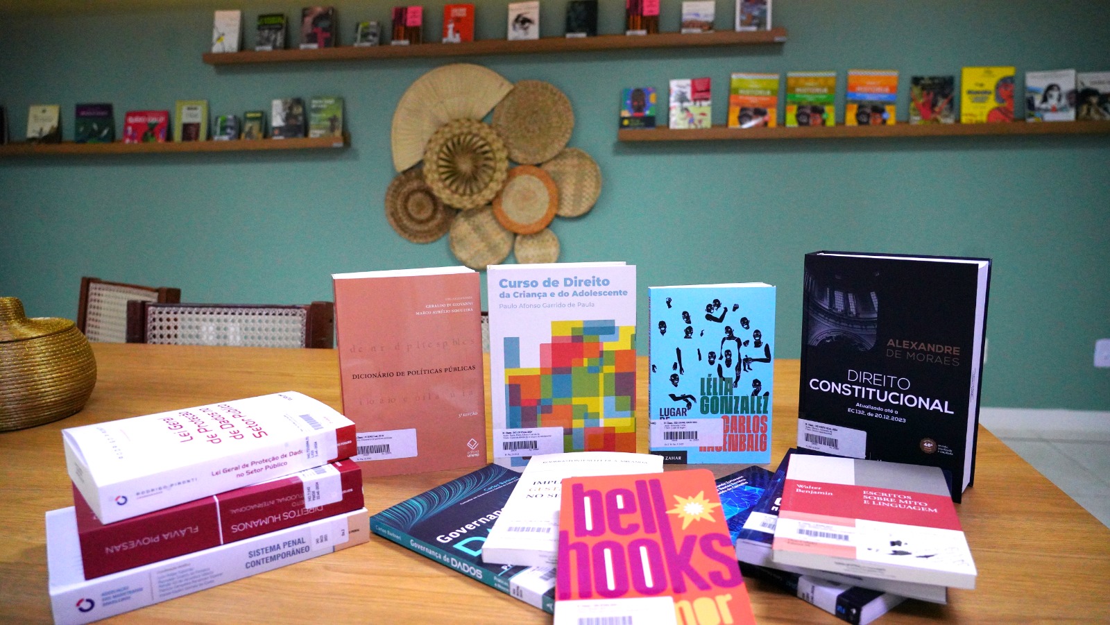 Seleção de livros em uma mesa, os livros estão organizados, alguns em pé e outros deitados em pilhas. Ao fundo, mais livros organizados em prateleiras.