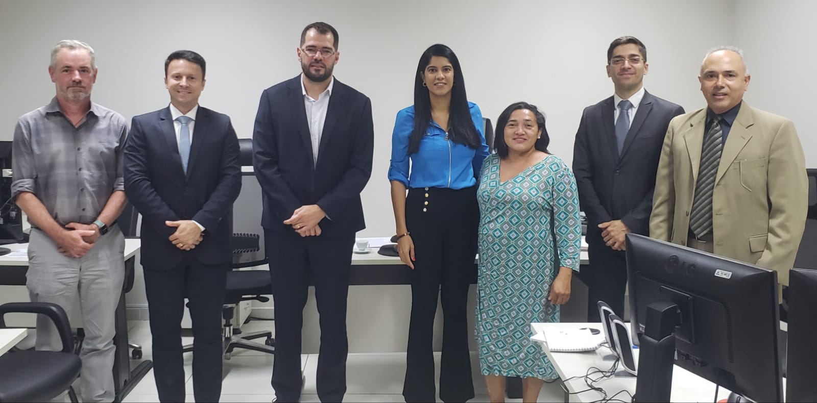 Pessoas posam para foto, duas mulheres de cabelos pretos com estaturas diferentes, uma menor e a outra maior, ao lado delas homens de diferentes idades usando ternos pretos, apenas um veste um terno verde claro, e um apenas com camisa social cinza