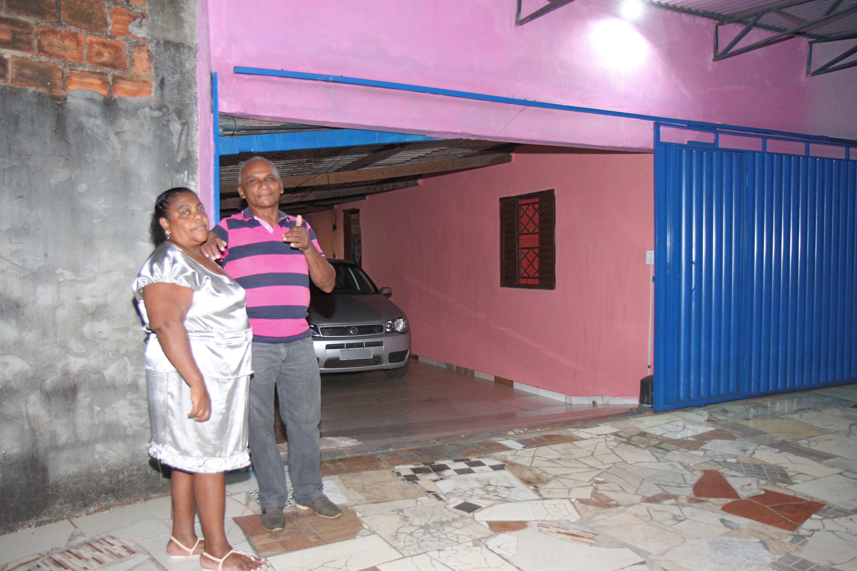 Eva Francisca de vestido dourado ao lado de seu marido de blusa rosa e listras azuis em frente a sua casa rosa e portão azul