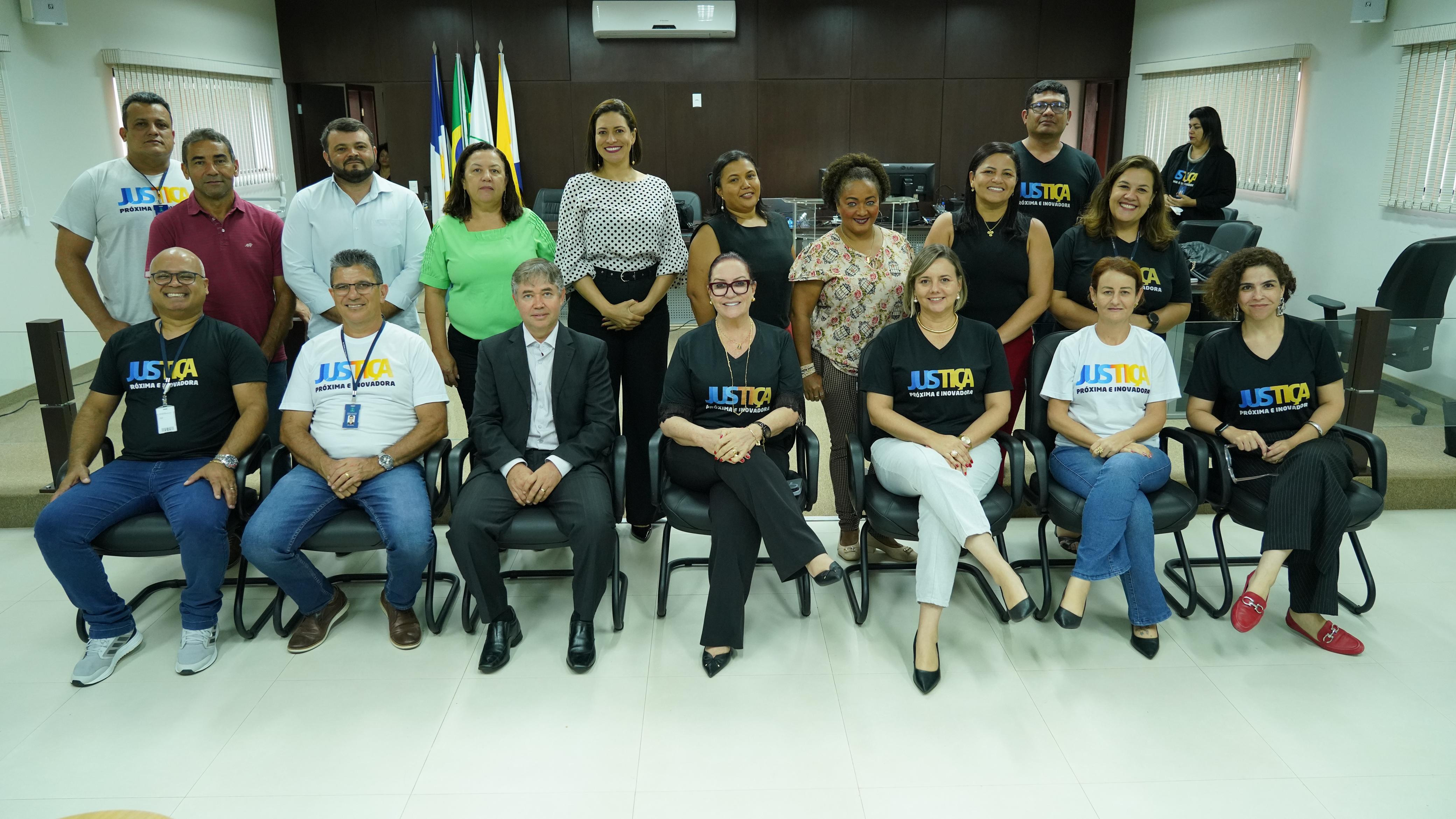 A imagem mostra um grupo de pessoas organizadas em duas fileiras em um ambiente de sala de reuniões. Na primeira fileira, estão sentadas mulheres e homens, vestindo camisetas pretas com a palavra "JUSTIÇA". Na segunda fileira, estão de pé mais pessoas, incluindo mulheres e homens, alguns usando camisas formais. Ao fundo, há bandeiras e o ambiente é institucional.