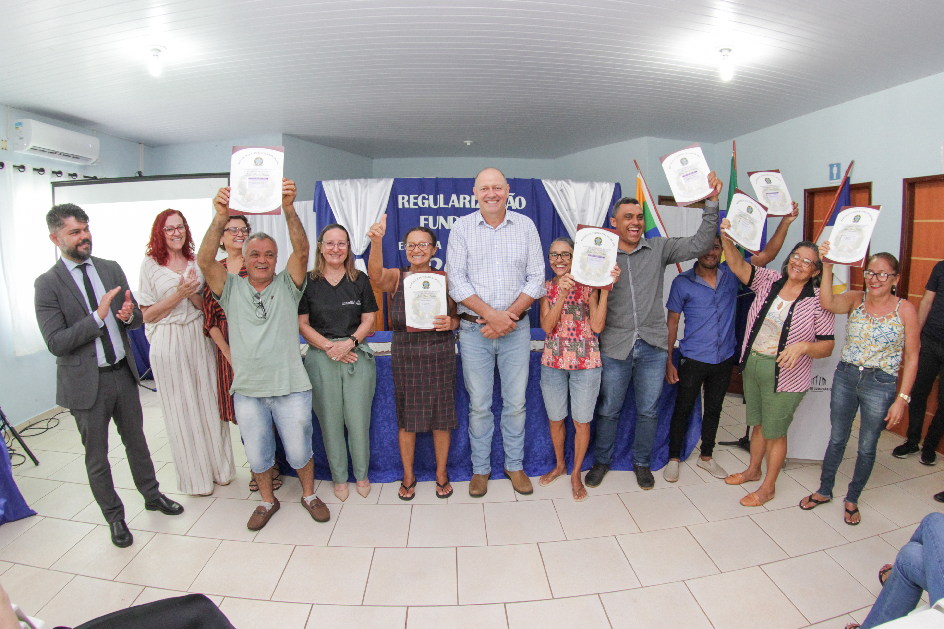 Pessoas posam para foto em sala com piso branco e paredes brancas, algumas delas erguem um documento