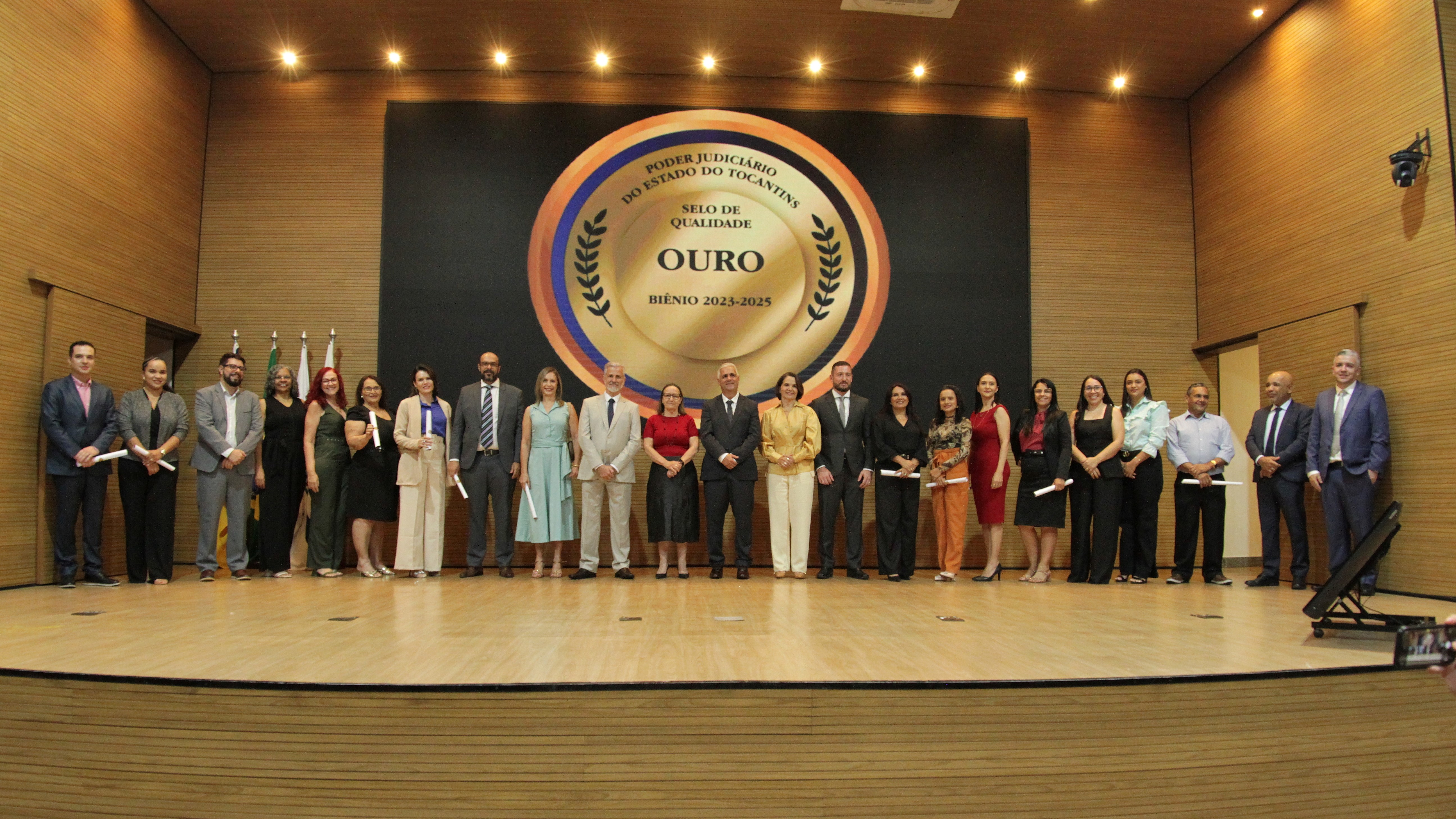 Grupo de pessoas em pé, alinhadas em um palco. No fundo há um painel circular e algumas pessoas estão segurando certificados.