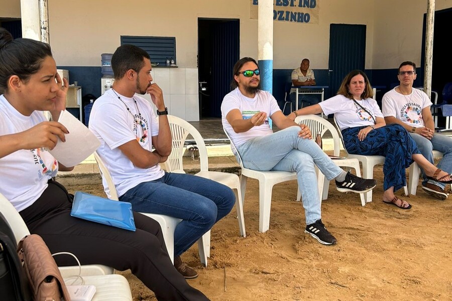 Na fotografia tem dois homens e duas mulheres com camisetas brancas, sentadas em cadeiras de plástico brancas e ao fundo tem um homem de pele negra e camiseta polo, observando 