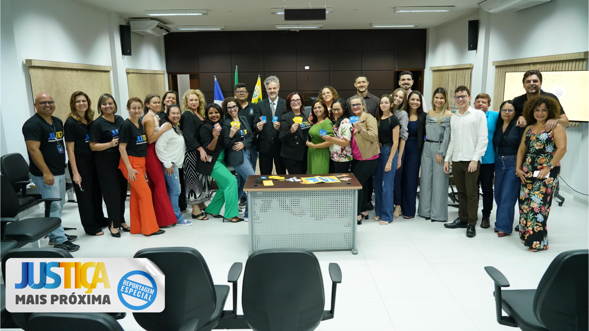 A imagem mostra um grande grupo de pessoas, com a maioria sendo mulheres, reunidas em uma sala de reuniões. Elas estão sorrindo e segurando pequenos cartões.  Há bandeiras e uma mesa com mais cartões no centro e tem  um logo de “Justiça Mais Próxima” na parte inferior da foto, ao redor tem algumas cadeiras vazias na cor preta.