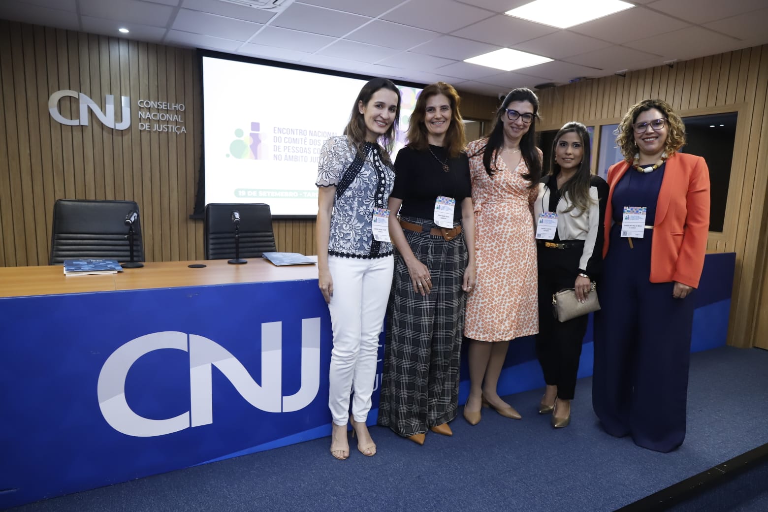 A imagem mostra cinco mulheres em pé em frente a uma mesa com um letreiro azul escrito "CNJ" com uma placa atrás delas com o texto "Encontro Nacional do Comitê dos Direitos das Pessoas com Deficiência no Âmbito do Judiciário". As mulheres estão vestidas de forma formal. Todas estão sorrindo para a câmera.