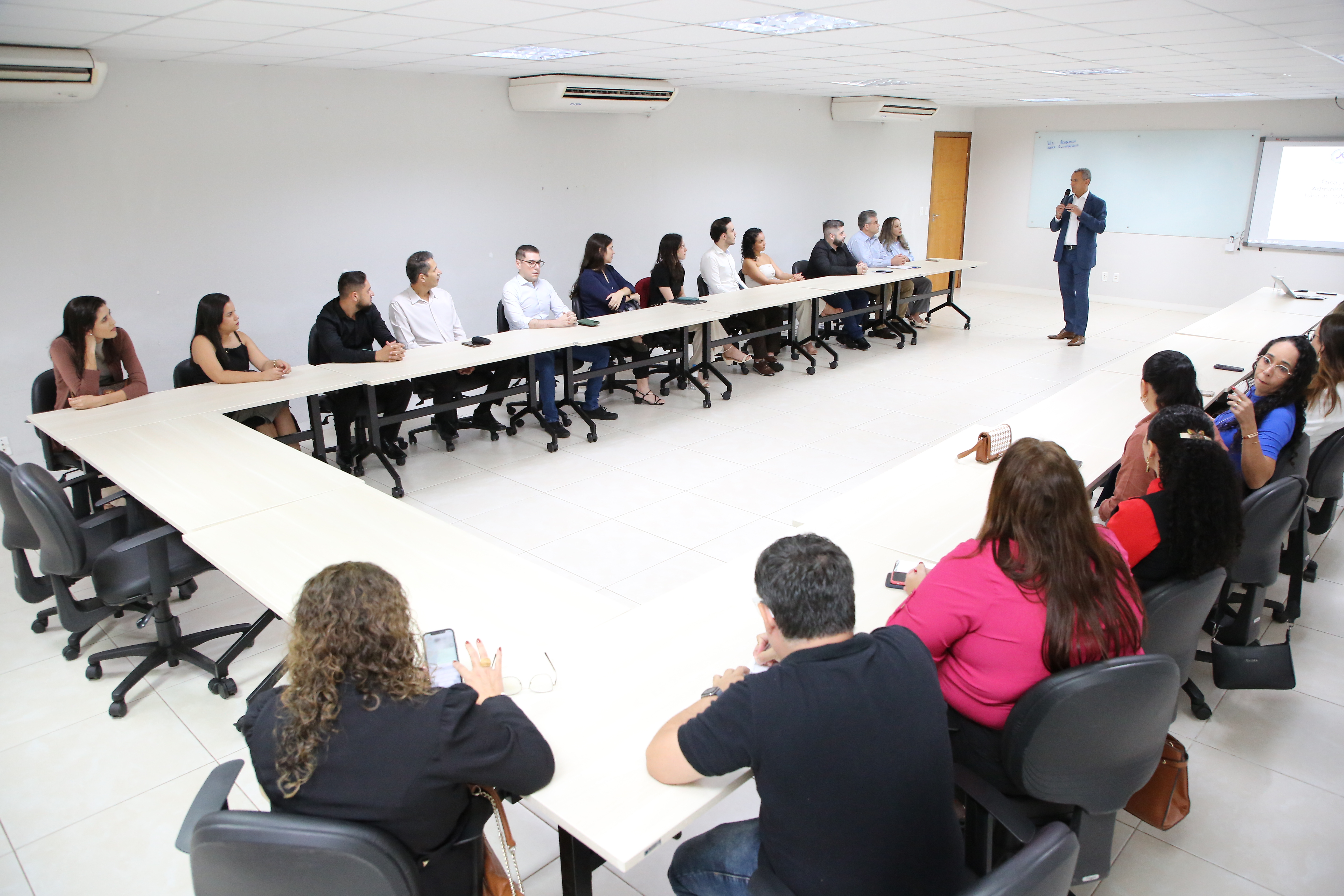 Homem em pé falando com um grupo de pessoas que estão sentadas. Na sala há mesas claras e um ambiente iluminado.