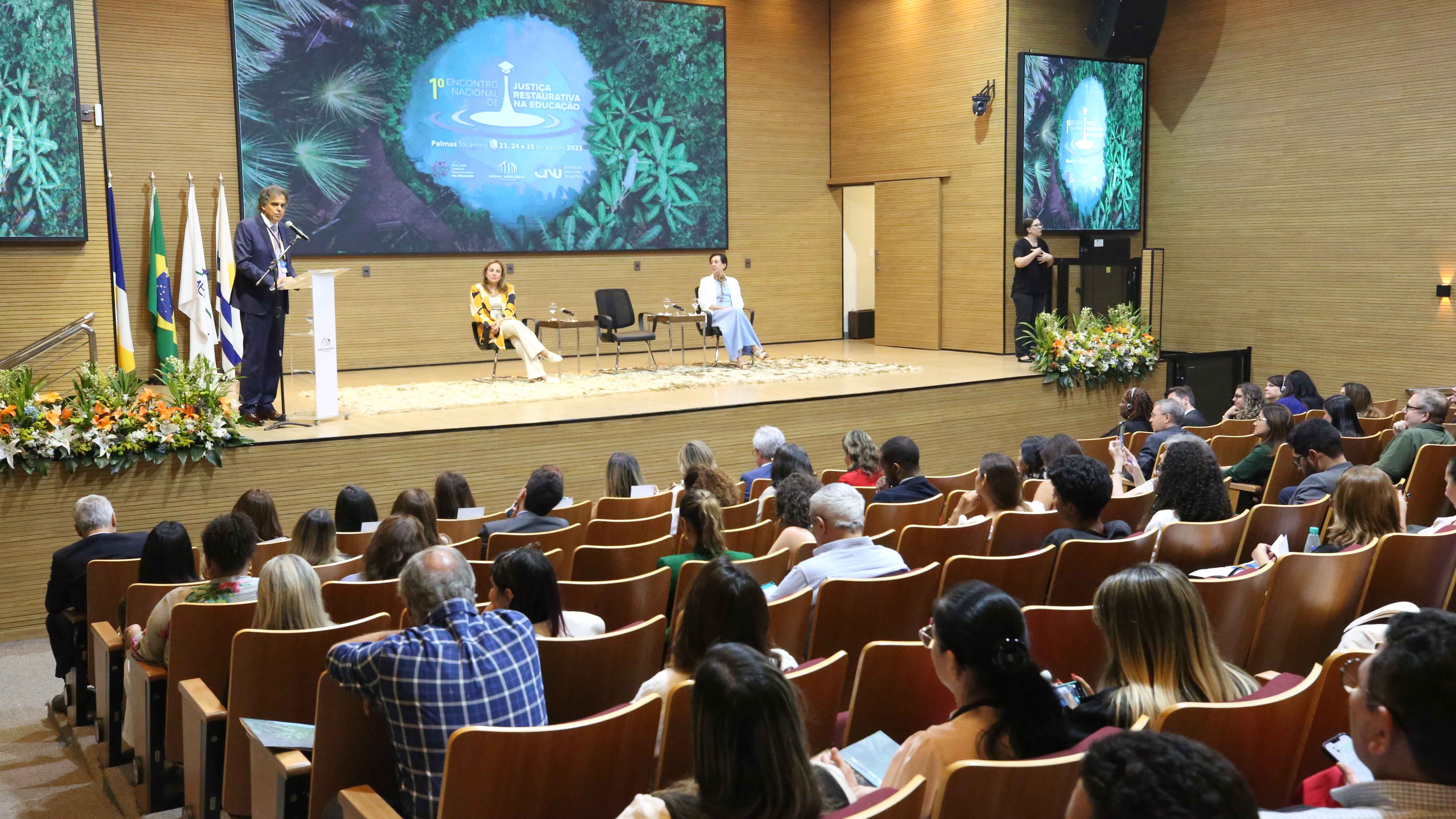 Ministro Vieira de melo fala ao microfone, observado pelas desembargadoras Angela Prudente e Angela Haonat, que estaão sentadas ao fundo, e pela plateia do auditório