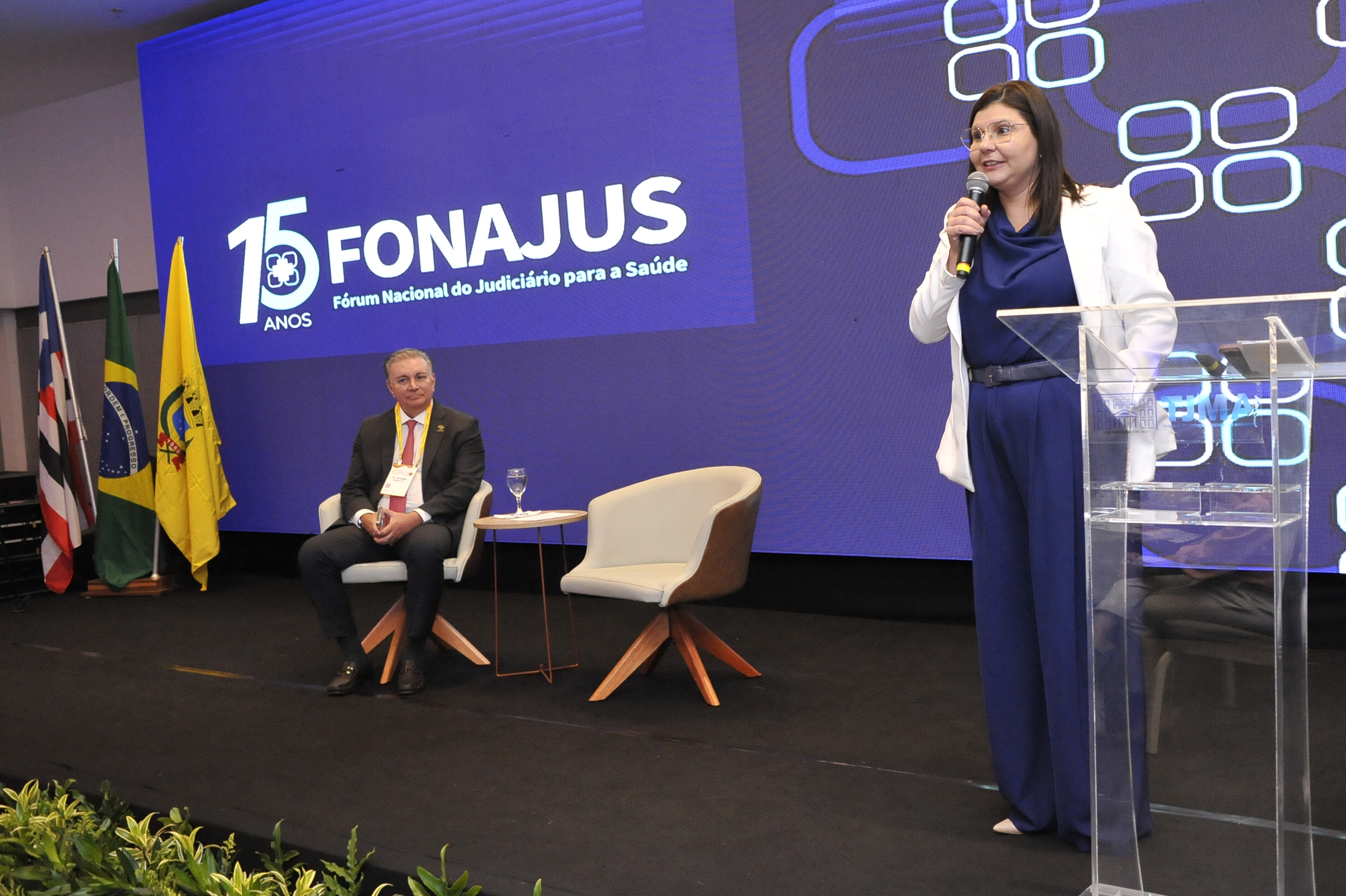 No centro, há um grande painel azul com as palavras "15 FONAJUS" e "Fórum Nacional do Judiciário para a Saúde". Uma mulher está em pé em um púlpito, vestindo um blazer branco e um macacão azul, segurando um microfone e falando. Ela usa óculos e parece concentrada. Ao lado dela, um homem está sentado em uma cadeira clara, vestindo um terno escuro e uma gravata vermelha, olhando para a mulher que fala. Entre eles, há uma mesa pequena com um copo de água. Atrás do homem, estão três bandeiras: a bandeira do Brasil e outras duas bandeiras, sendo uma delas amarela. A iluminação é clara e destaca os participantes do evento.