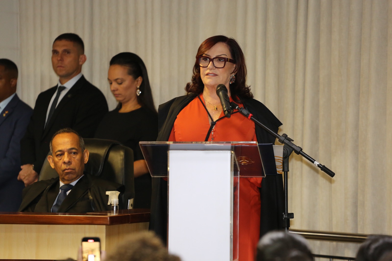 Desembargadora Etelvina, de vestido laranja e beca preta por cima, falando ao microfone