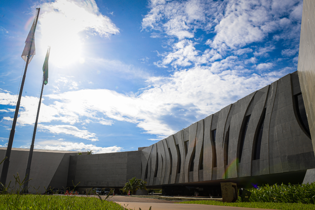 Fachada cinza com detalhes em cimeto, céu azul com bastante nuvens, grama verde que fica em volta de uma passarela de cimento.