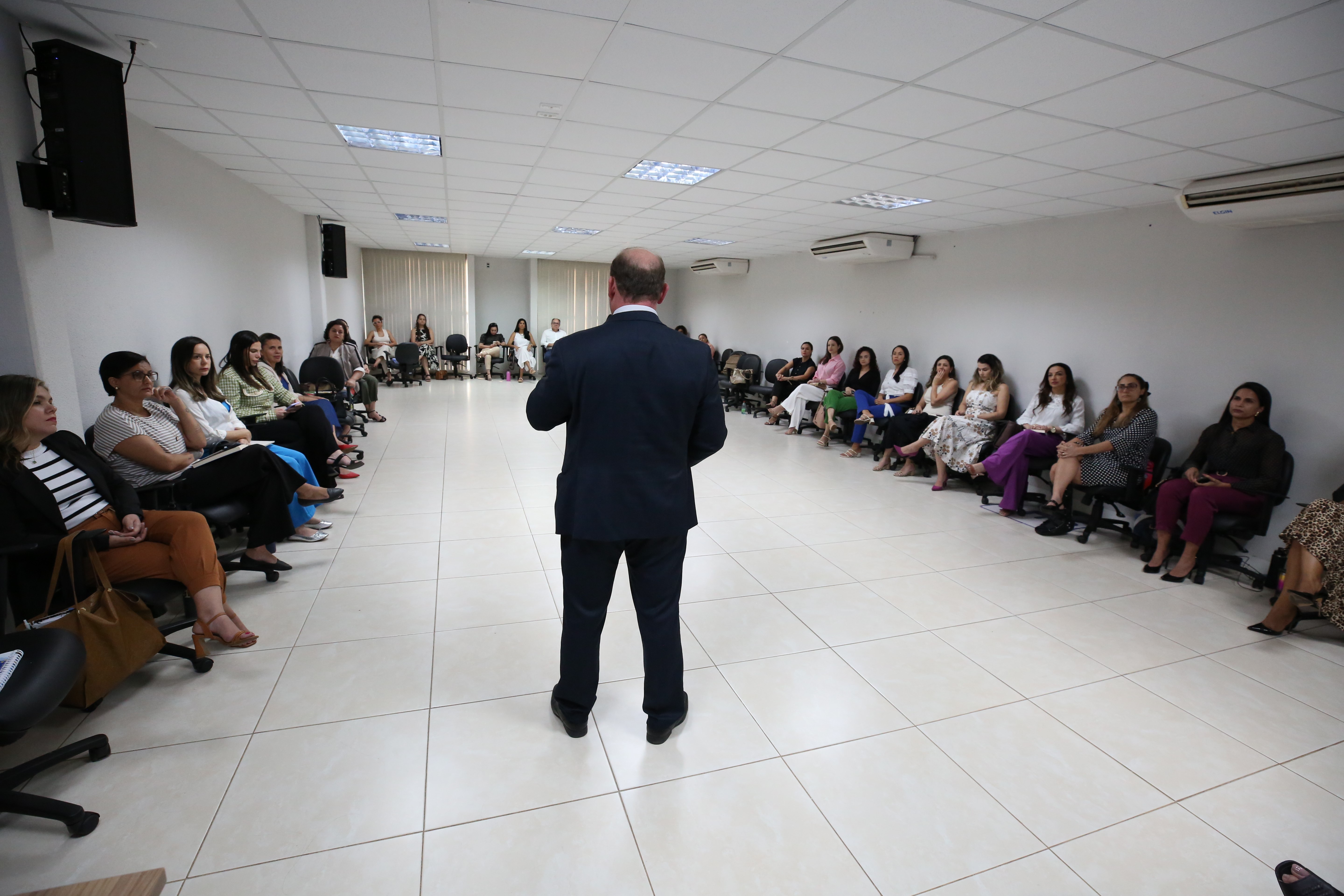 Homem de terno está em pé, falando para um grupo de pessoas sentadas em uma sala ampla.