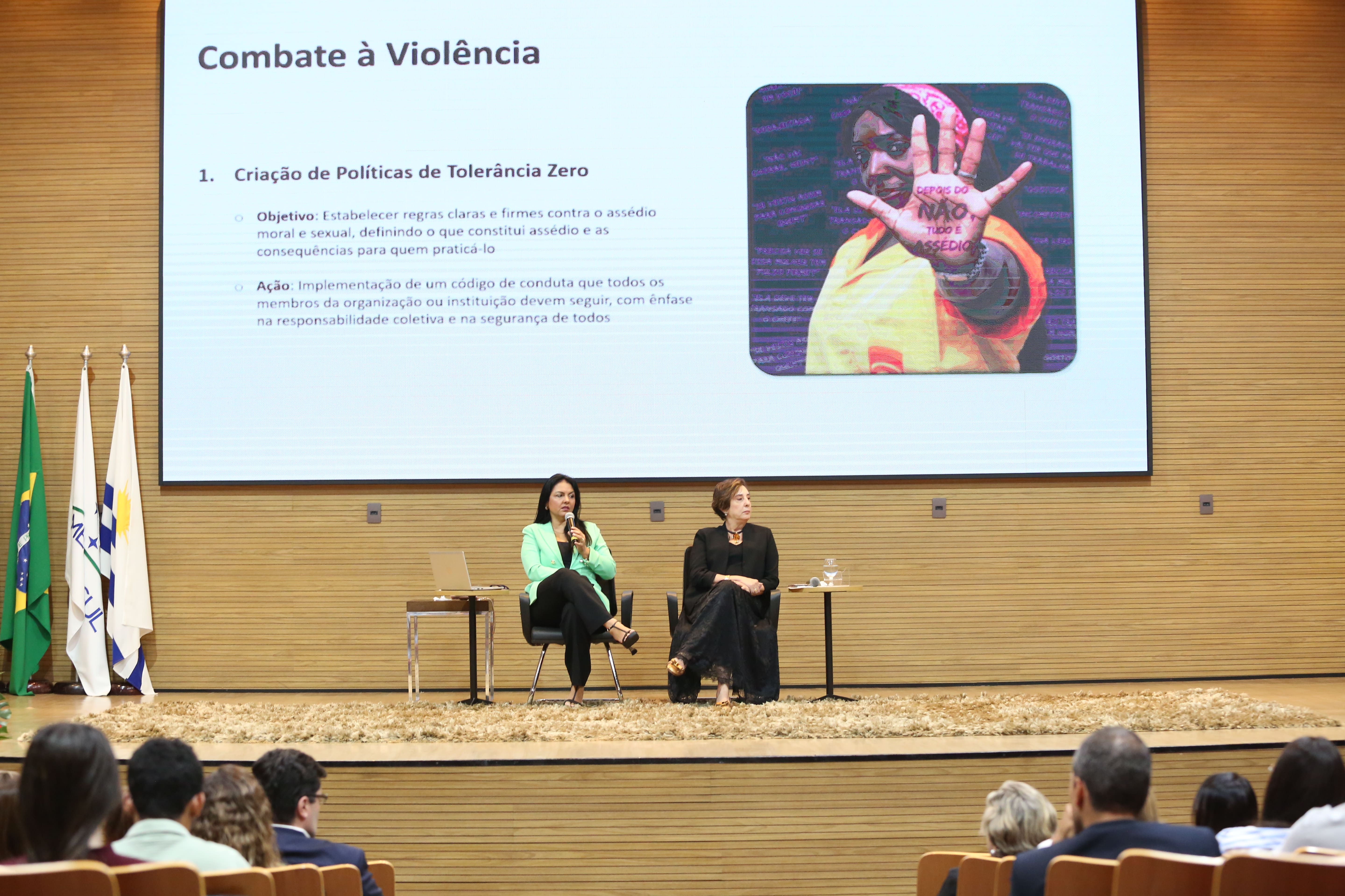 Duas mulheres com trajes formais em uma sala bem iluminada com um telão ao fundo com os dizeres "combate a violência".