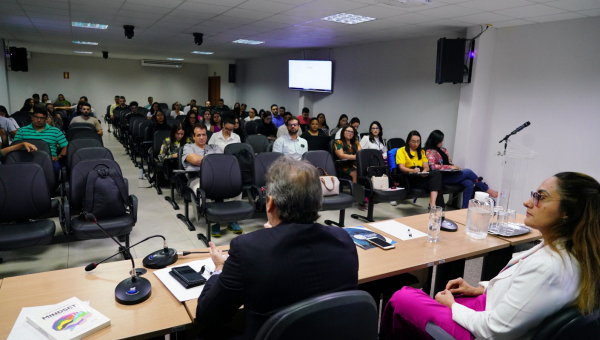 Sala de reunião com pessoas sentadas em cadeiras que estão em fileiras e duas pessoas a frente, um homem e uma mulher.