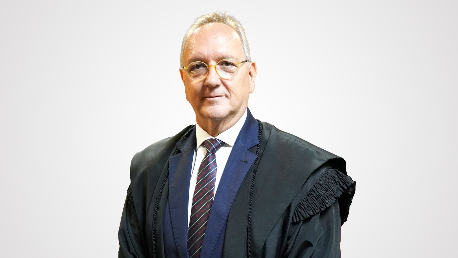 desembargador Pedro Nelson, homem branco de cabelos brancos; ele usa óculos e veste camisa branca, gravata listrada com detalhes em preto, cinza e vermelho, blazer azul escuro e toga preta por cima