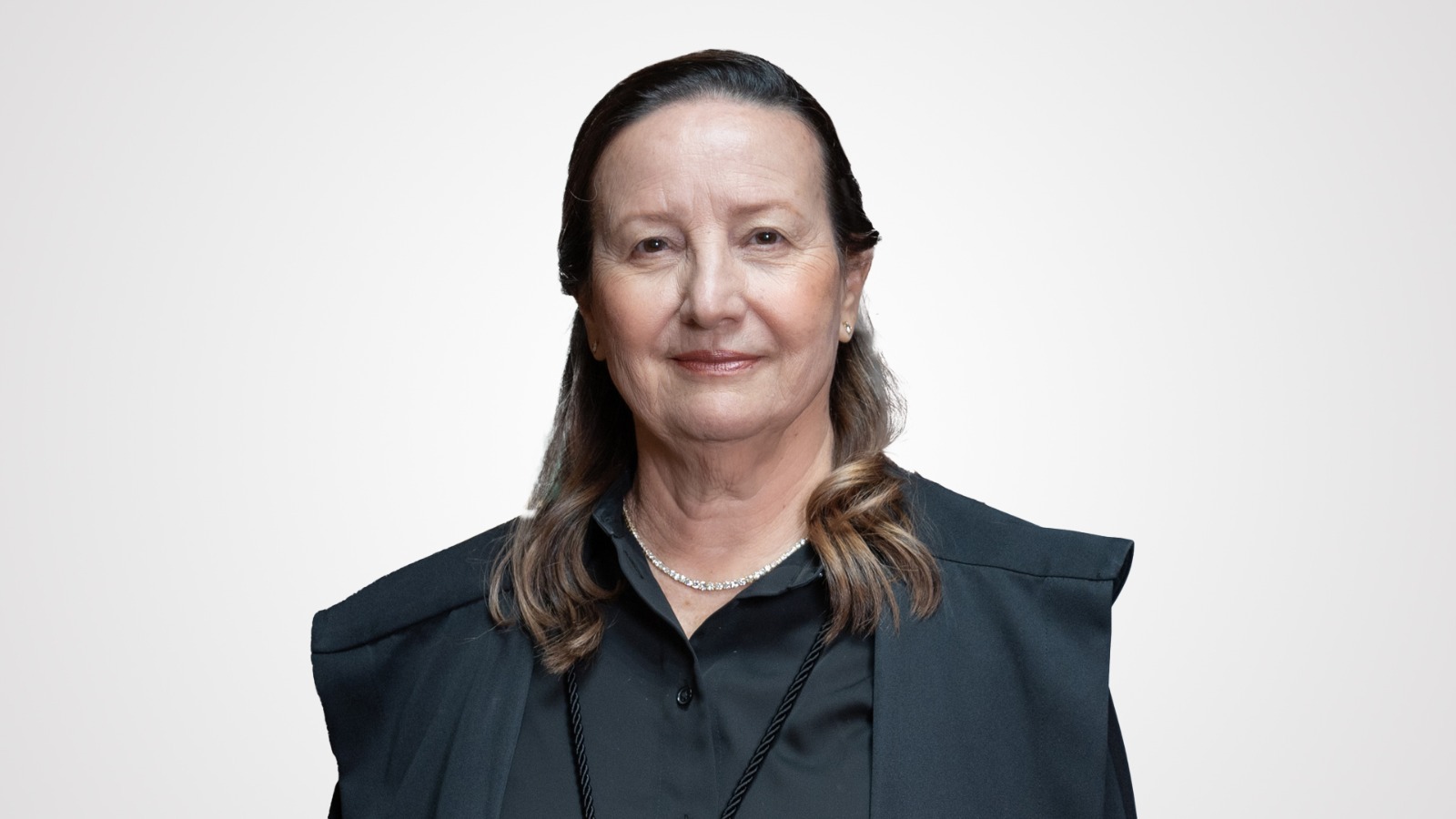 Desembargadora Maysa é uma mulher branca de cabelos castanhos ; ela usa roupa preta com uma toga escura por cima; está em pé, com as mãos cruzadas na altura da cintura, à frente das bandeiras do Tocantins, Brasil, Mercosul e Palmas 