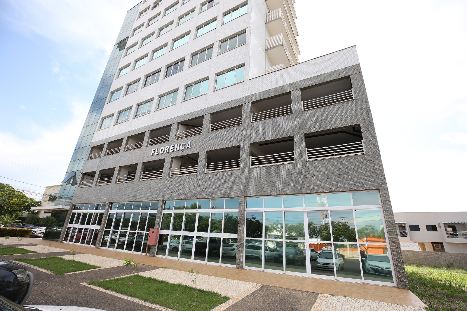 Prédio moderno com fachada de vidro e tijolo, com o nome "FLORENÇA" em letras grandes na fachada. O prédio tem várias janelas e um estacionamento com carros estacionados. Na frente do prédio, há um gramado verde e uma calçada. O céu está azul com algumas nuvens brancas.