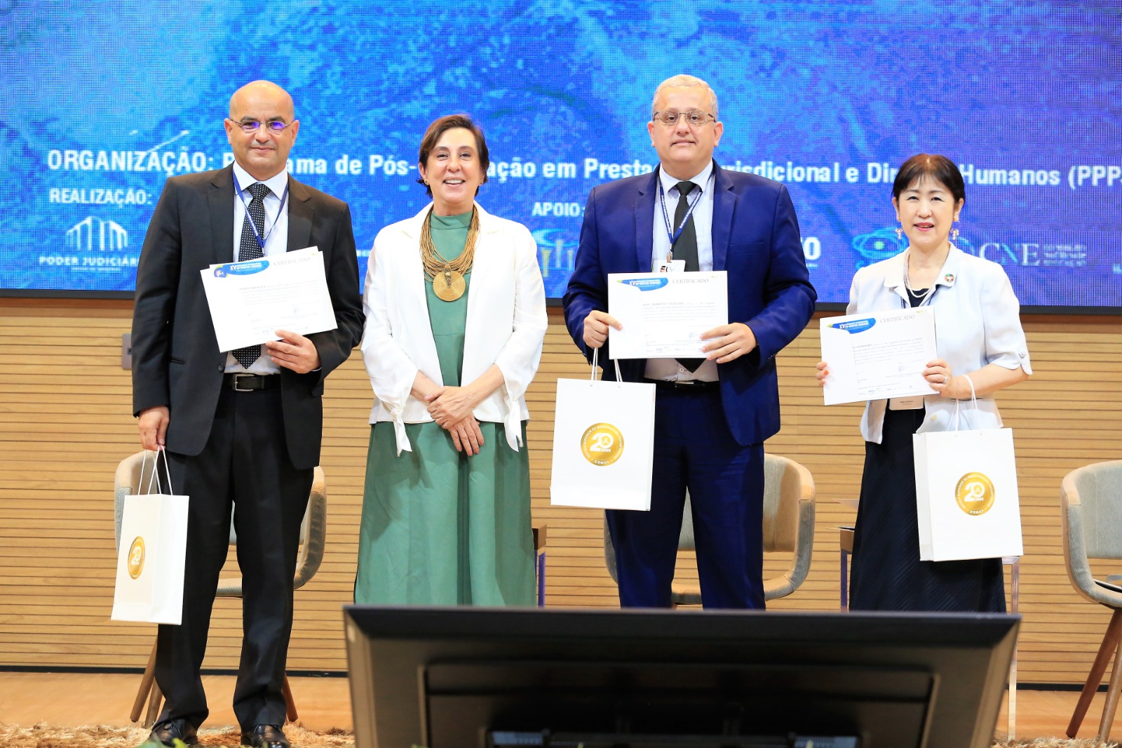 Quatro pessoas no pauco segurando certificados e sacolas; são duas mulheres e dois homens