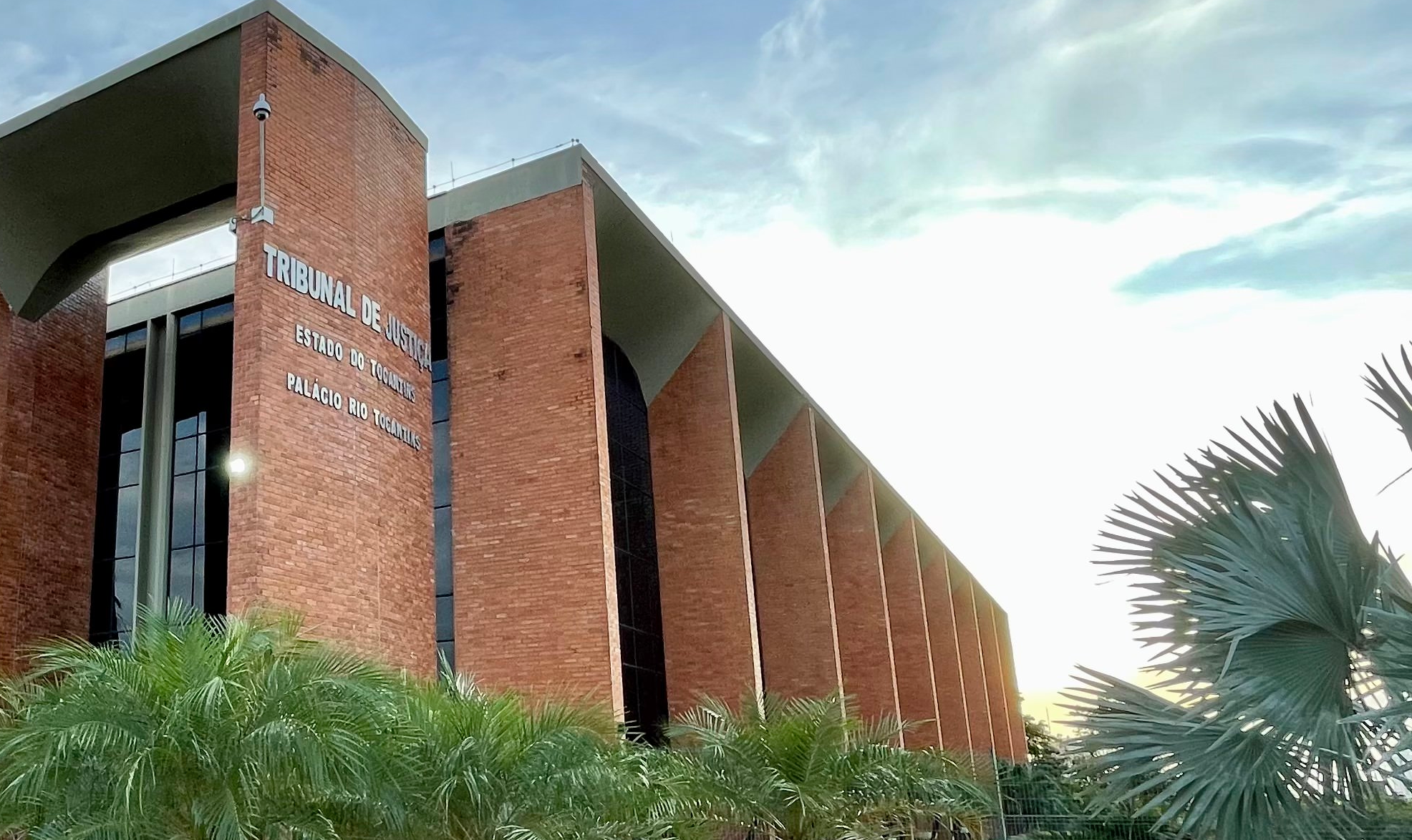 A imagem mostra a fachada do Tribunal de Justiça do Estado do Tocantins (Palácio Rio Tocantins), com arquitetura imponente em tijolos aparentes e grandes pilares verticais. O prédio é cercado por áreas ajardinadas, com vegetação ornamental.