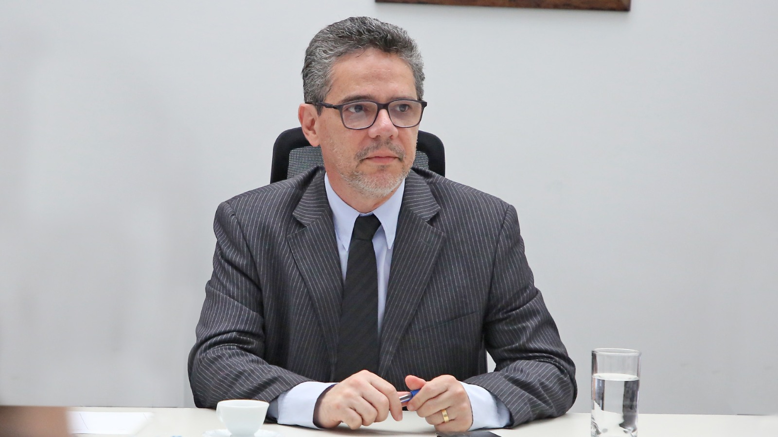 Juiz Adriano Gomes de Melo Oliveira sentado, usando óculos, terno cinza escuro, camiza azul claro e gravata preta