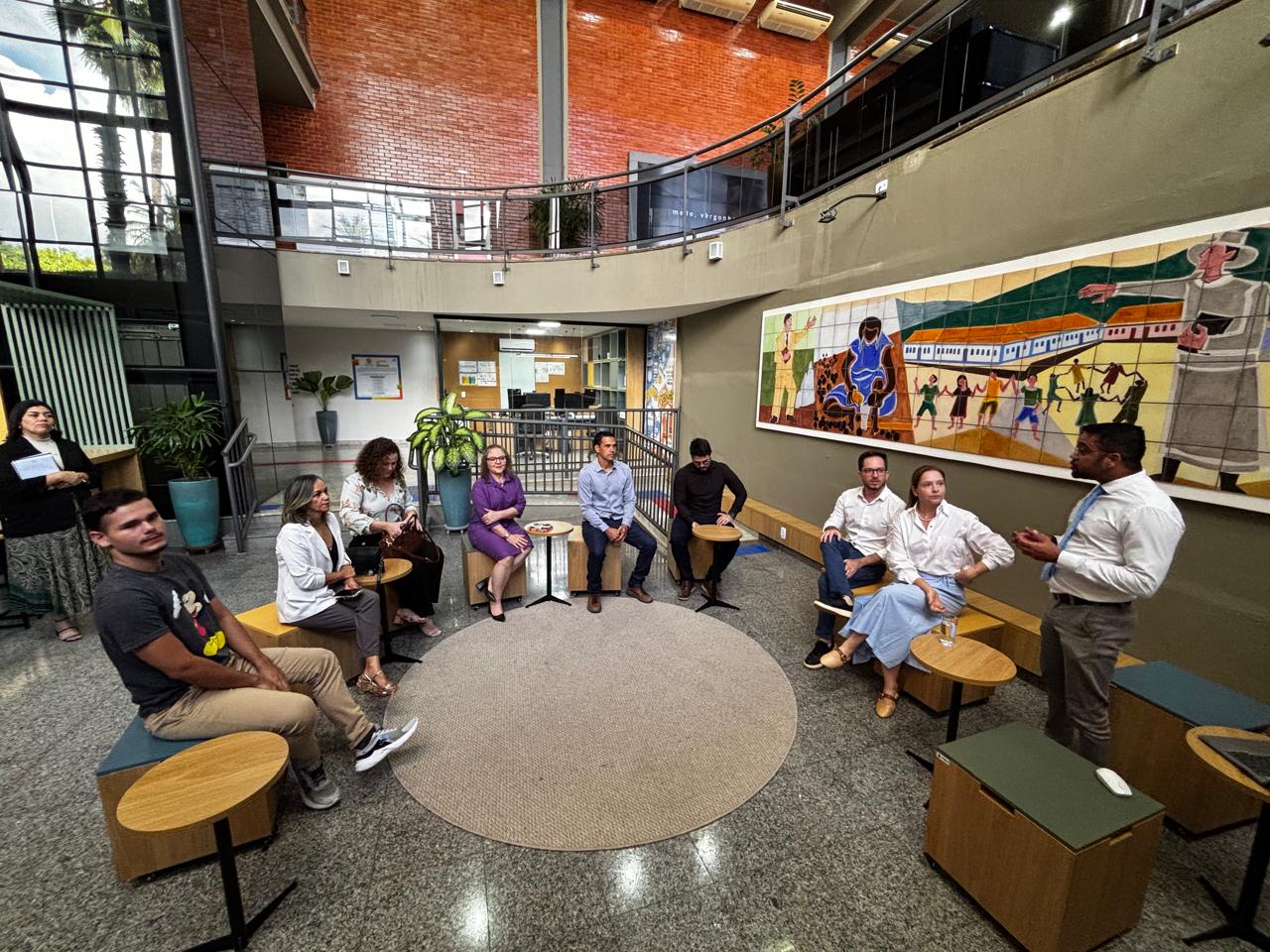 Homens e mulheres sentados em bancos de madeira instalados em espaço arejado dentro do TJTO onde funciona o laboratório de inovação. Homem negro de camisa branca e calça bege apresenta o espaço aos presentes. Nas paredes cinzas laterais, há dois painéis coloridos, obras de artistas