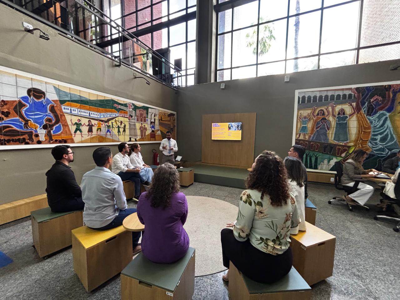 Homens e mulheres sentados em bancos de madeira instalados em espaço arejado dentro do TJTO onde funciona o laboratório de inovação. Homem negro de camisa branca e calça bege apresenta o espaço aos presentes. Nas paredes cinzas laterais, há dois painéis coloridos, obras de artistas