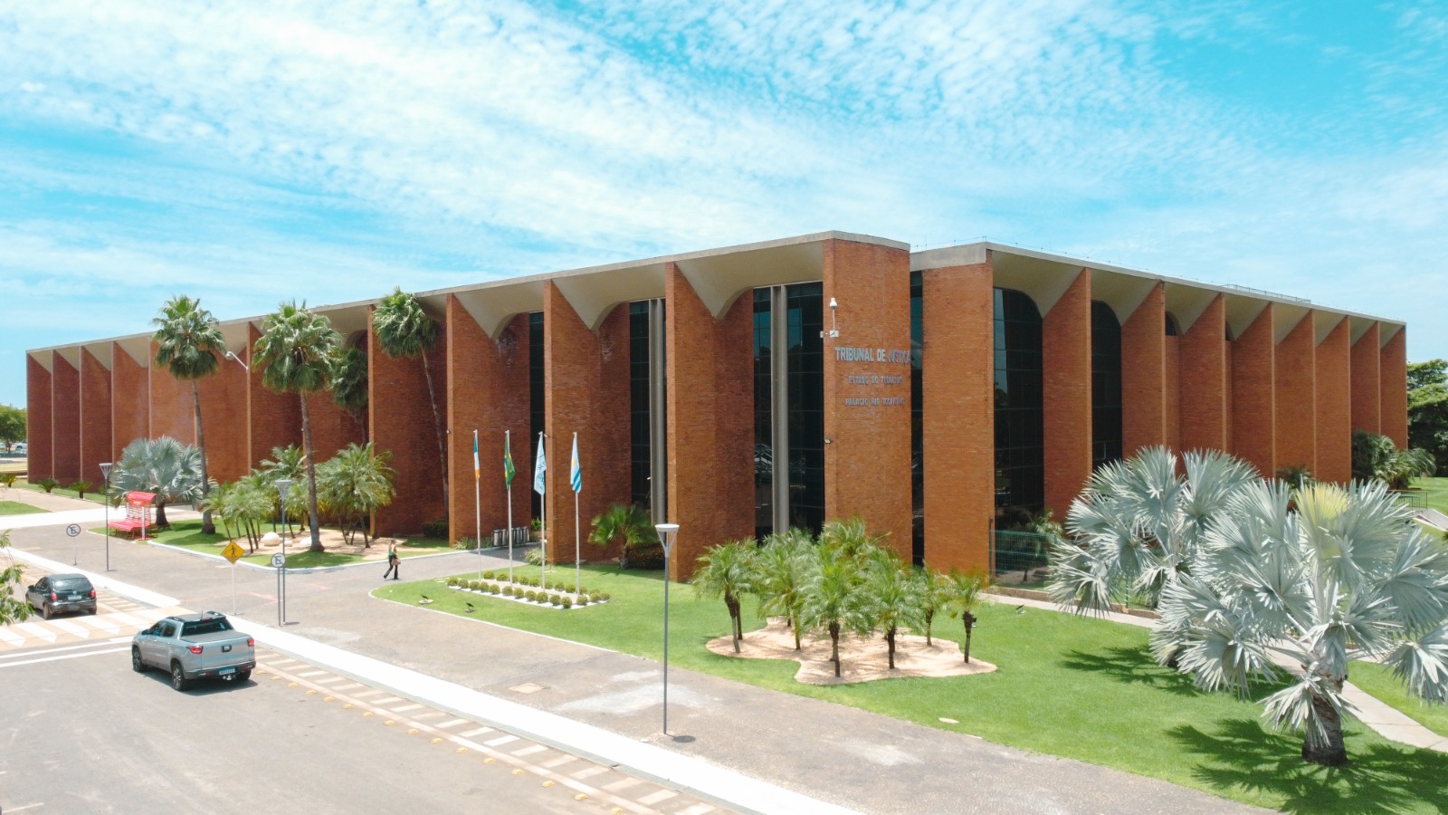 Prédio do Tribunal de Justiça do Tocantins, com fachada em tijolos aparentes, grandes colunas e painéis de vidro. À frente, jardim com palmeiras, mastros com bandeiras e via de acesso com veículos, sob céu azul.