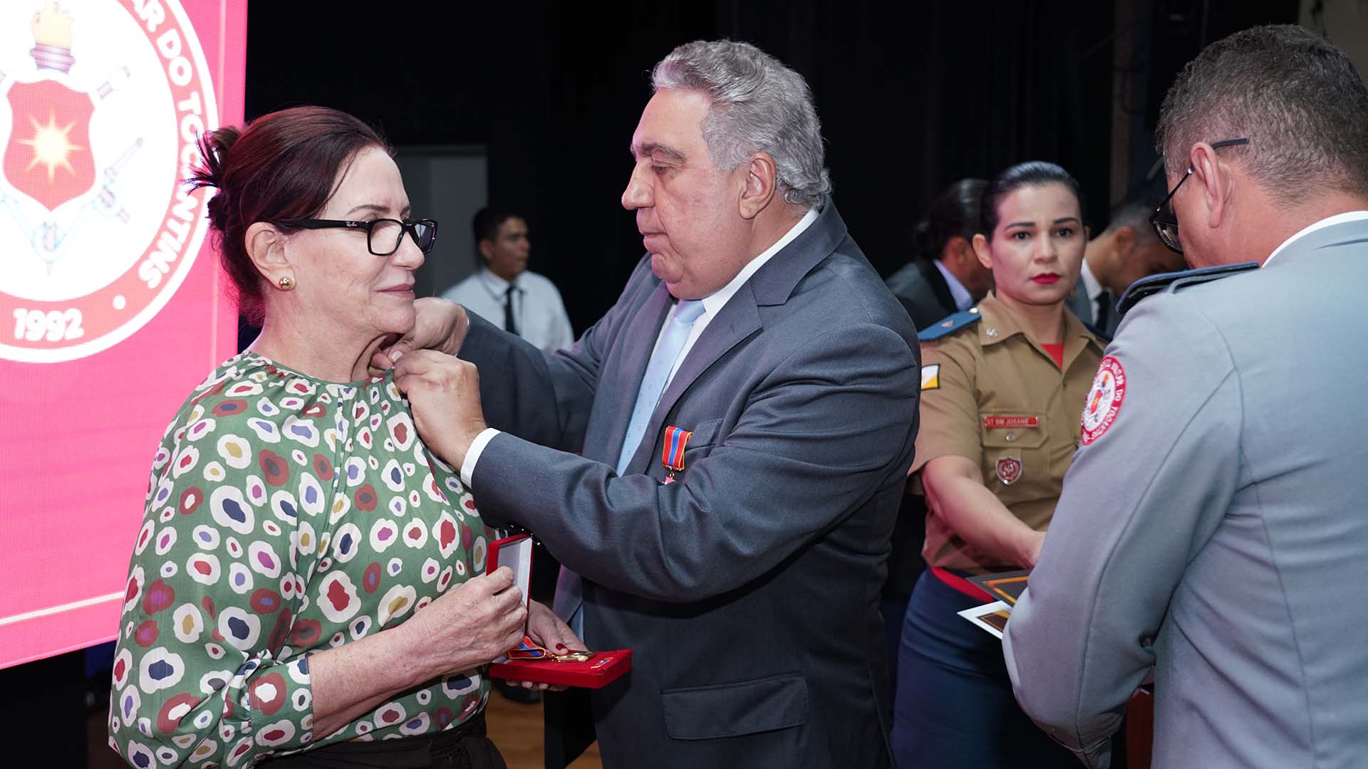 desembargadora Etelvina, de cabelos presos, óculos e blusa estampada em tons de verde, branco e vermelho, recebendo a honraria do vice-governador Laurez Moreira