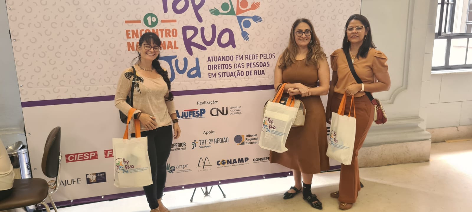 Juíza Rosa Maria, de roupa marrom e sapato preto mais ao centro, com duas servidoras do TJTO ao seu lado; todas usam óculos e estão com bolsas e segurando sacolas do evento; a servidora da direita usa uma blusa e calça em tons terroso e a da esquerda usa blusa bege e calça preta; ao fundo um grande painel com as informações do evento