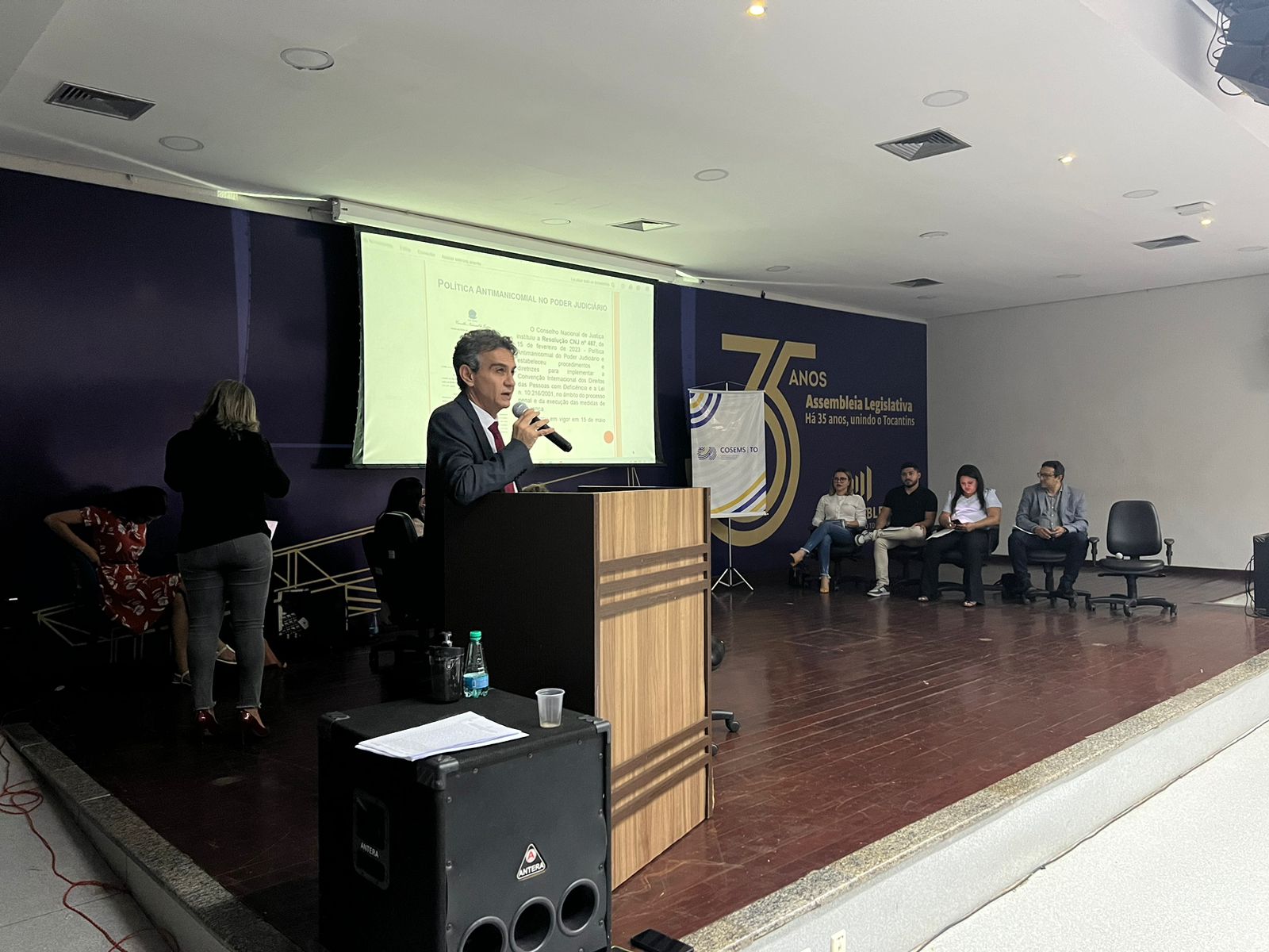 Homem de cabelos grisalhos em pé, ao púlpito, com microfone na mão, falando para público de auditório. Ao fundo, telão com apresentação no meio de uma parede azul escura