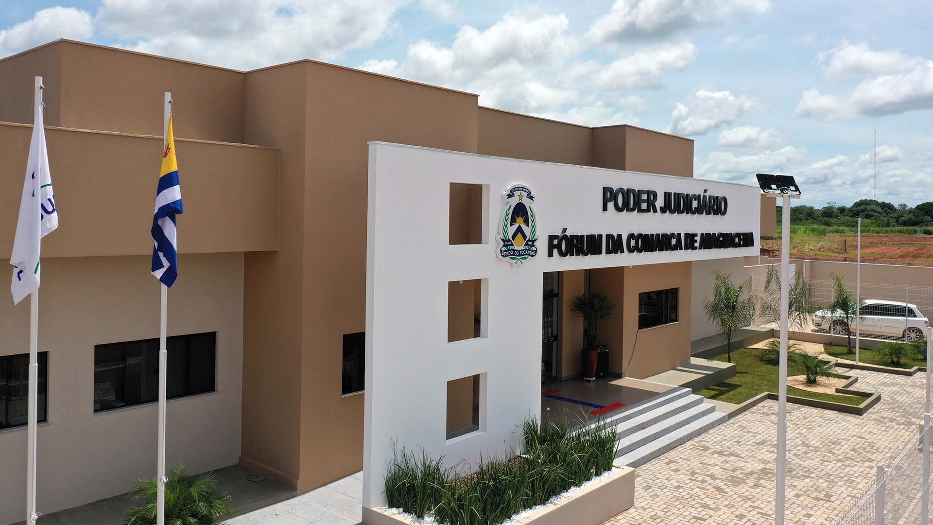 Fachada do Fórum de Araguacema vista da perspectiva lateral, com parte do estacionamento, jardim frontal e o pórtico branco com letreiros na cor preta