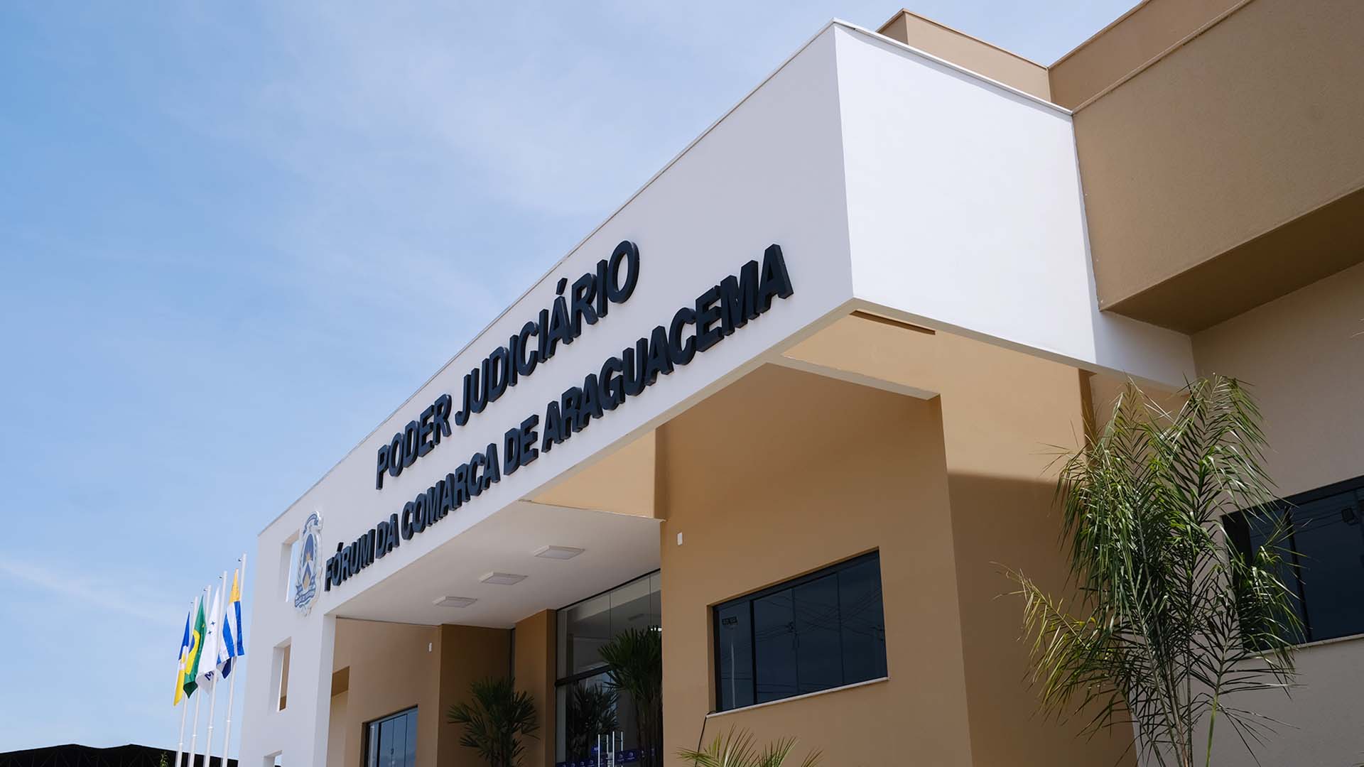 Fachada do prédio do Fórum de Araguacema, da perspectiva lateral, com vista do pórtico branco com letreiros na cor preta, parte das paredes marrons e mastros com bandeiras