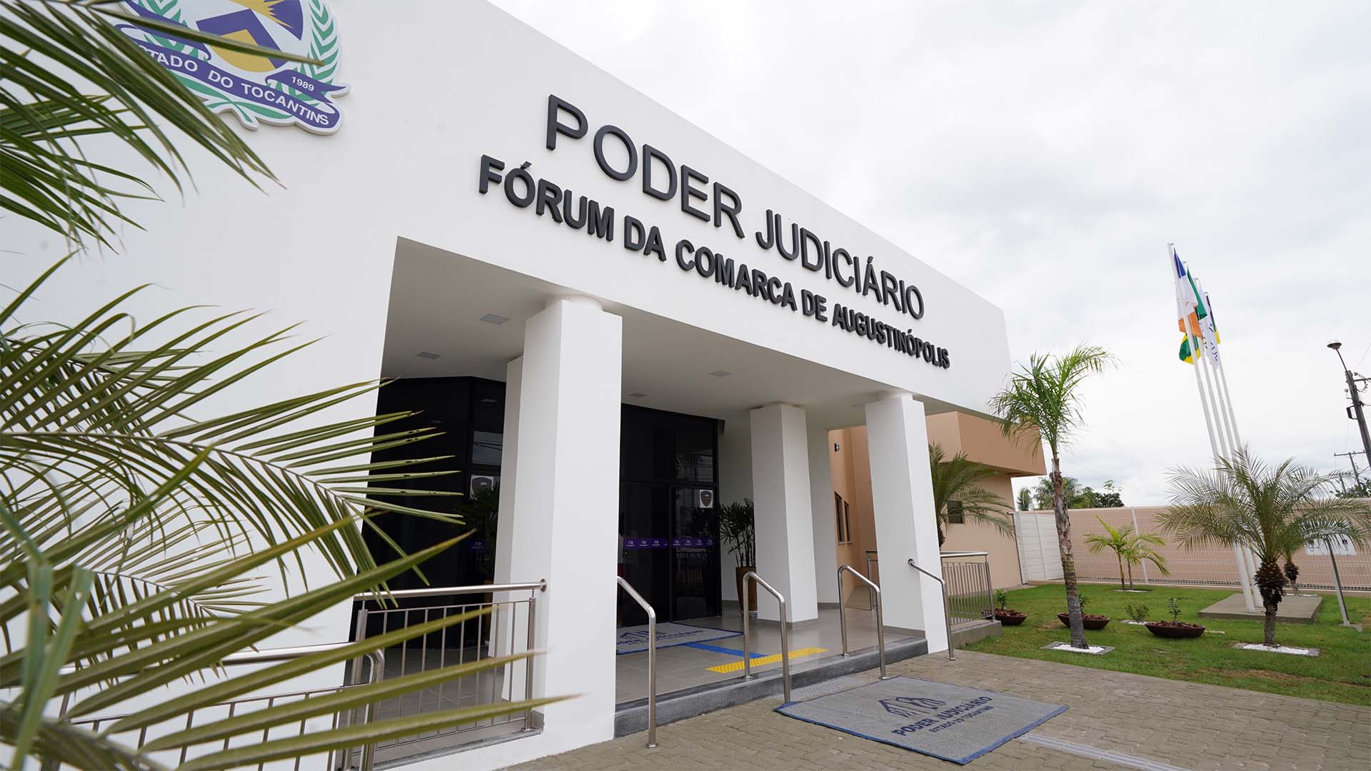 Fachada do prédio da Comarca de Augustinópolis. Estrutura conta com paredes brancas. portas e janelas de vidro escuro, degraus e corrimãos. Na Frente a logo e o nome: Poder Judiciário - Fórum da Comarca de Augustinópolis