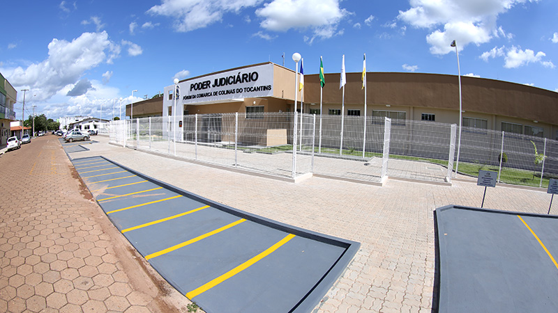 Fachada do Fórum de Colinas do Tocantins da perspectiva lateral, com parte do estacionamento, jardim frontal, janelas espelhadas e o pórtico branco com letreiros na cor preta
