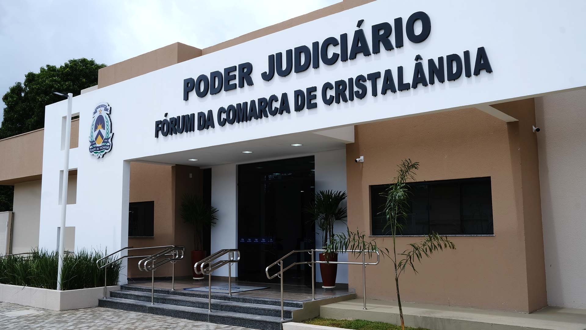 Fachada do Fórum da Comarca de Cristalândia, com letreiro “Poder Judiciário – Fórum da Comarca de Cristalândia” na parte superior e brasão do Estado do Tocantins à esquerda. Entrada com portas de vidro, escada e rampa com corrimãos metálicos. Paredes em tons de branco e bege e plantas ornamentais na área externa.