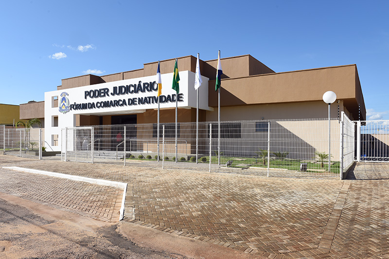 Fachada do prédio da Comarca de Natividade; paredes nas cores marrom, bege e branca; protas e janelas de vidro; à frente grades de proteção, jardim, luminárias e mastros com bandeiras.