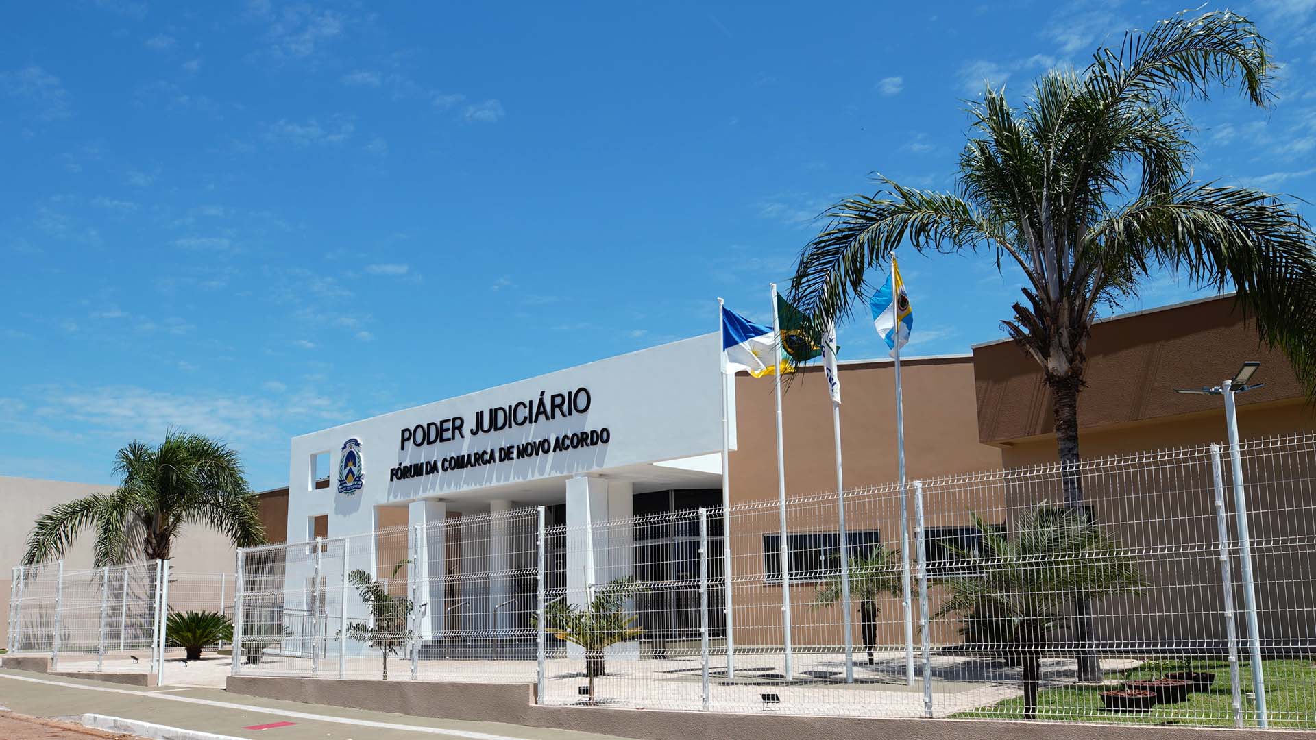 Fachada do fórum de Novo Acordo, visto de fora do alambrado de proteção, com pórtico branco e letreiro na cor preta, paredes marrons, jardim e coqueiro