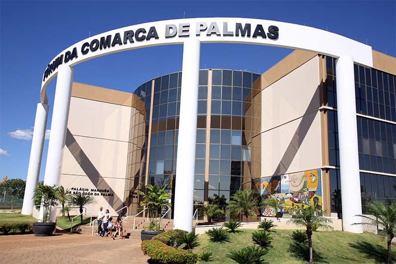 Fachada do prédio do Fórum da Comarca de Palmas, com paredes pintadas nas cores bege e marrom, vidraças espelhadas e arcos brancos à frente com o letreiro com o nome: Fórum da Comarca de Palmas; à frente um jardim com grama, plantas e palmeiras