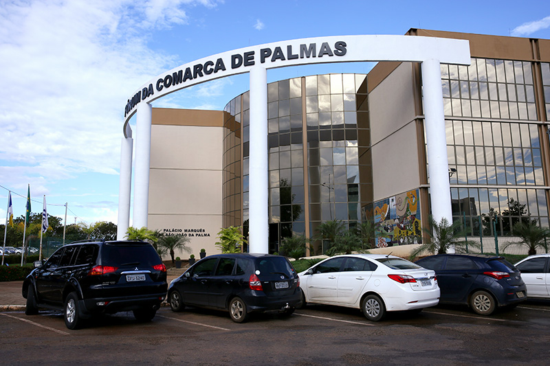 Foto mostra carros estacionados à frente do prédio do Fórum de Palmas. O prédio tem cor marrom e branco e tem vidros nas laterais