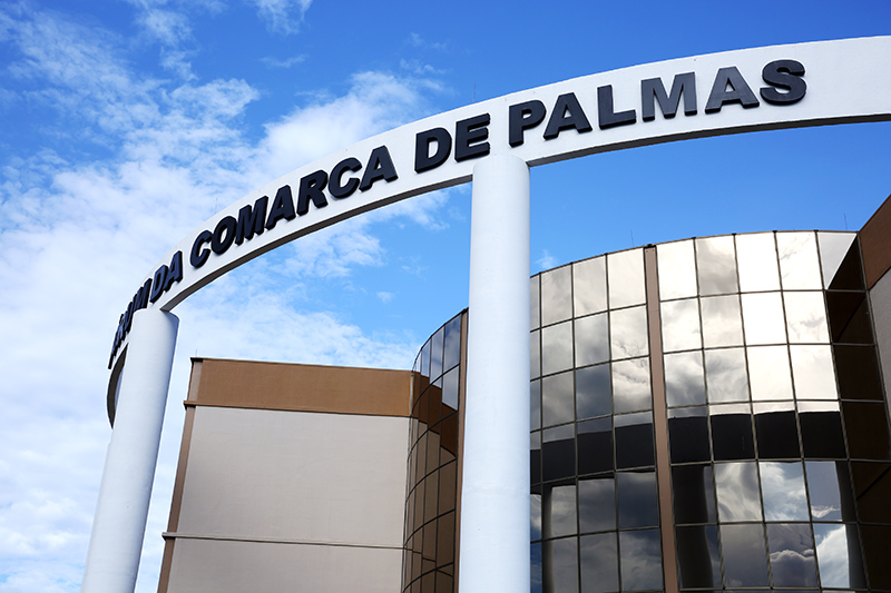 Foto de detalhe da Comarca de Palmas; na imagem aparecem parte das paredes e colunas pintadas nas cores bege, marrom e branca, vidraças e o arco frontal com o nome Comarca de Palmas com letras pretas