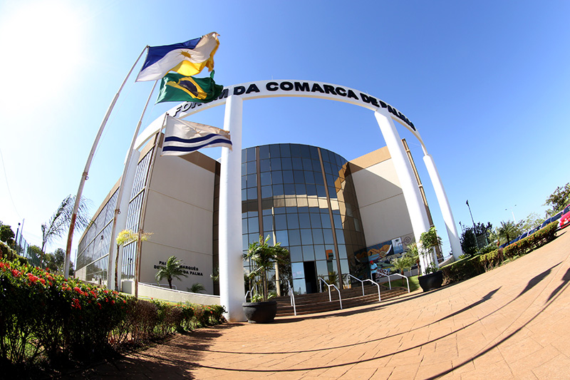 Fachada do Fórum de Palmas, vista em perspectiva frontal distorcida, mostrando a frente espelhada, mastros com bandeira à esquerda da imagem e o pórtico com letreiros na cor preta